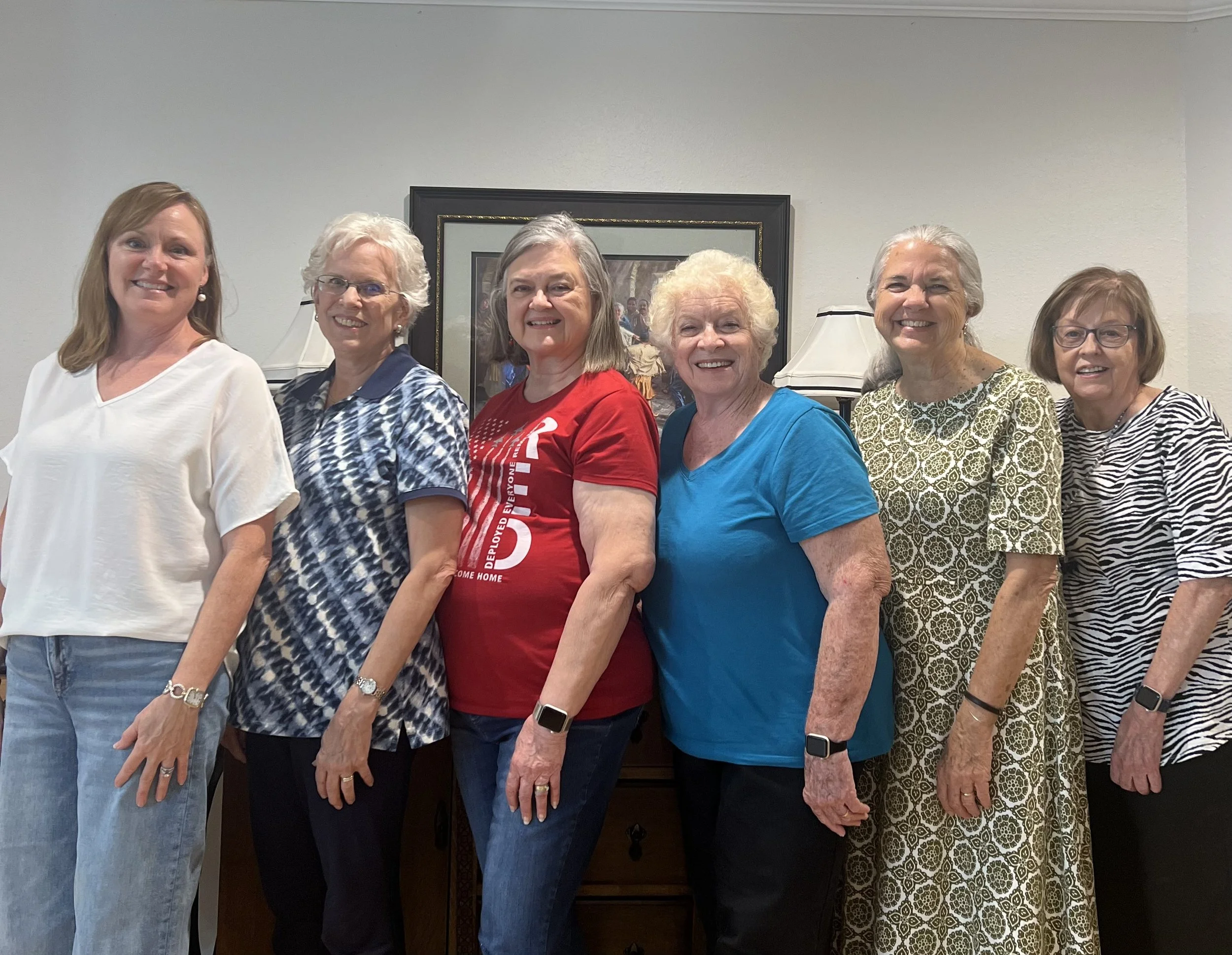 Seven women standing side by side indoors, smiling for the camera, with a framed picture and lamps in the background.
