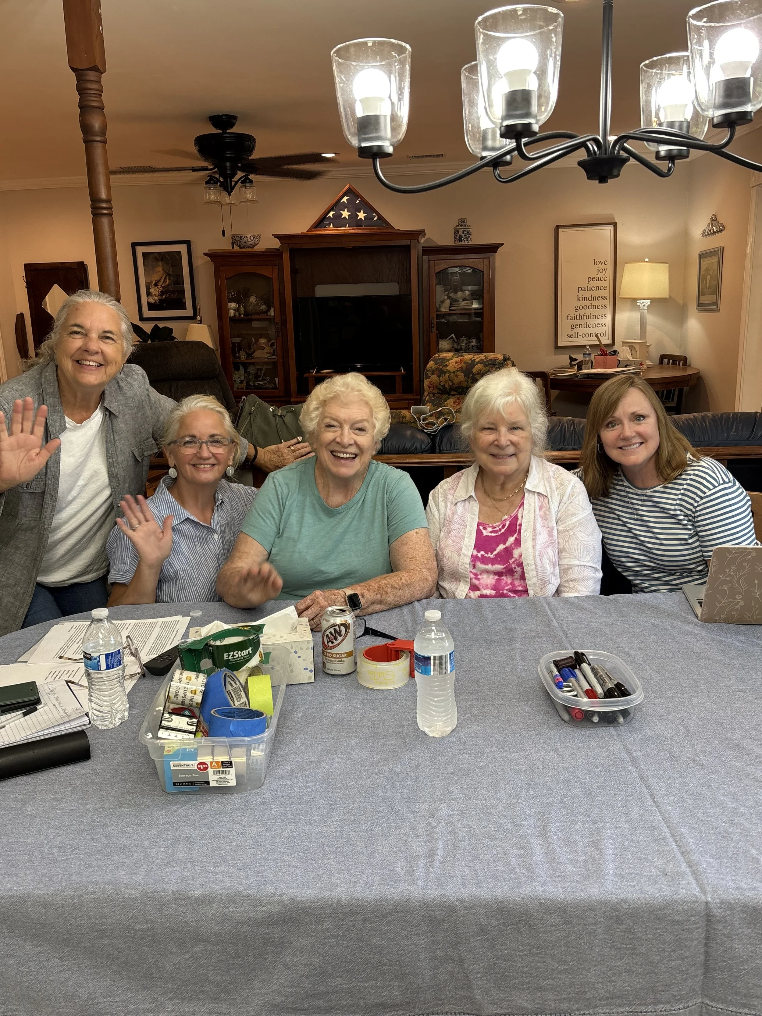 Group of five women sitting at a table in a cozy living room, smiling and waving at the camera, with various drinks and supplies on the table.