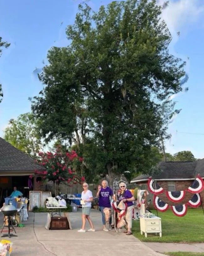 Three women standing outdoors in front of a large tree, with parked houses and items on tables around them, and patriotic bunting hanging on the right side of the scene.
