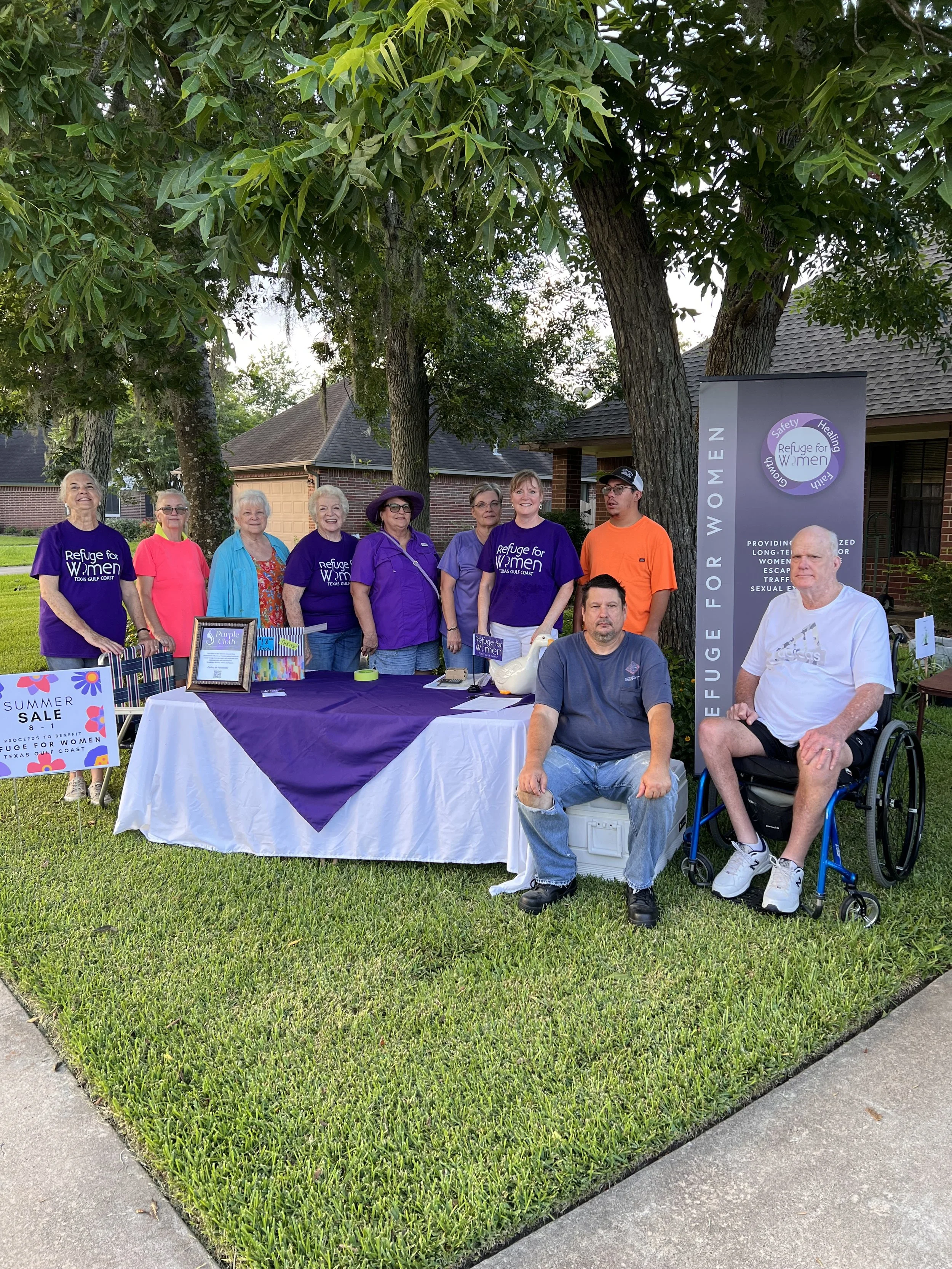 A group of people standing behind a table at an outdoor event under trees. Several women are wearing purple shirts with the logo 'Refuge for Women.' Two men are seated in front, one in a wheelchair and another on a cooler. The table has a purple cloth, framed pictures, and a sign that says 'Summer Sale.' There is a banner that reads 'Refuge for Women' with additional text about providing housing and support for women.