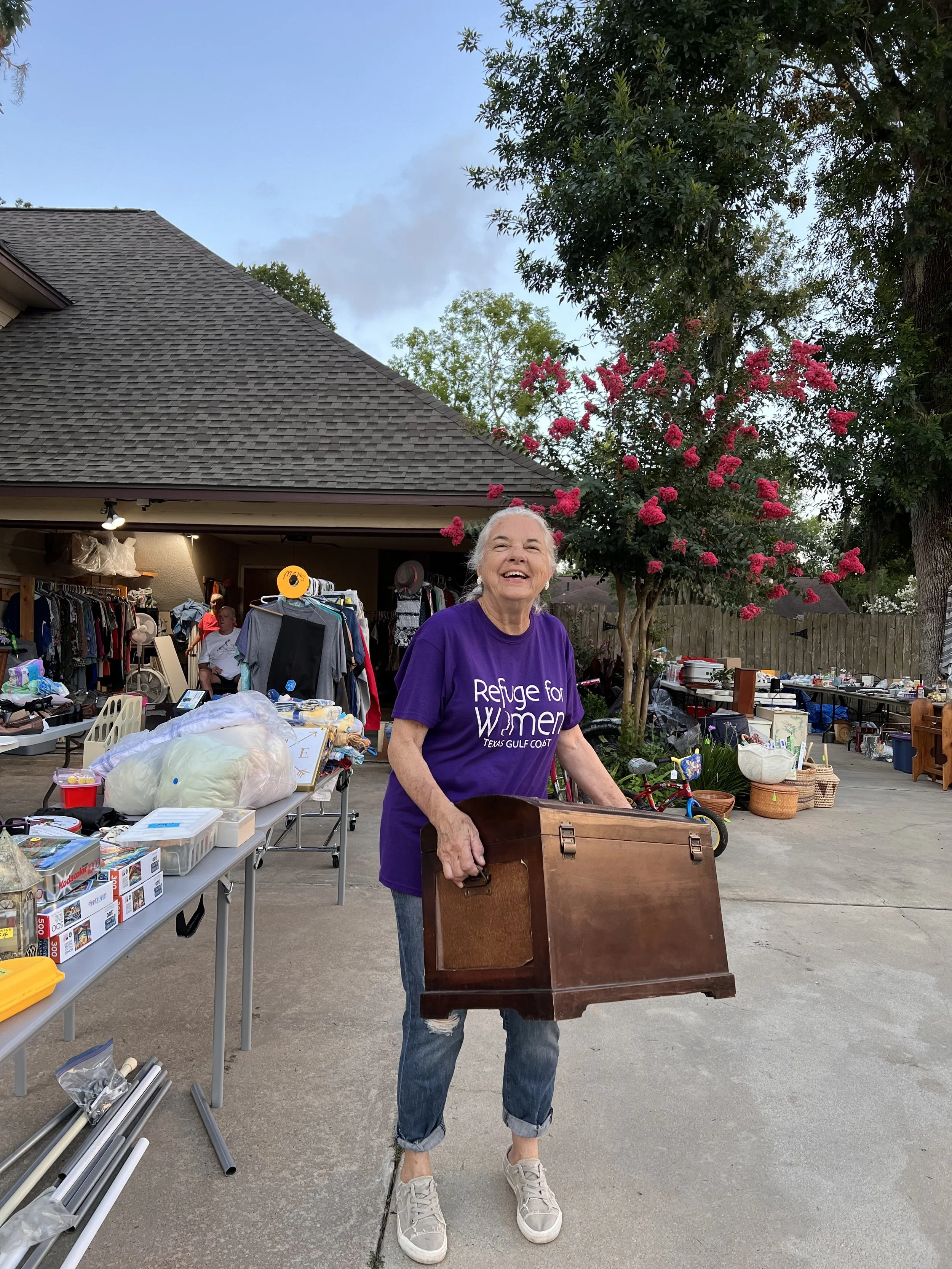 Older woman holding a wooden box at a yard sale in a residential neighborhood, smiling, with tables of miscellaneous items and clothing in the background, a pink flowering tree, and a house with a sloped roof and trees.