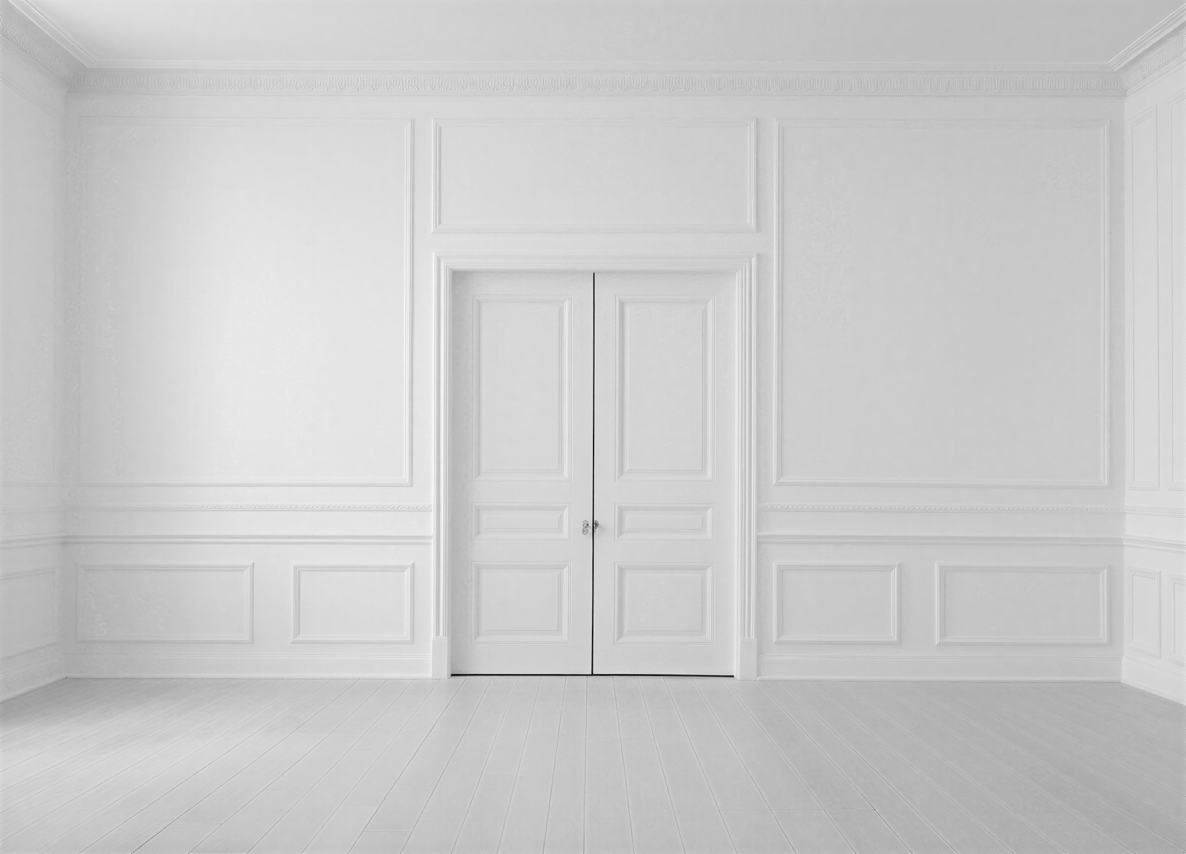 A room with white walls, white ceiling, decorative crown molding, and white wooden floor, featuring a white double door in the center.