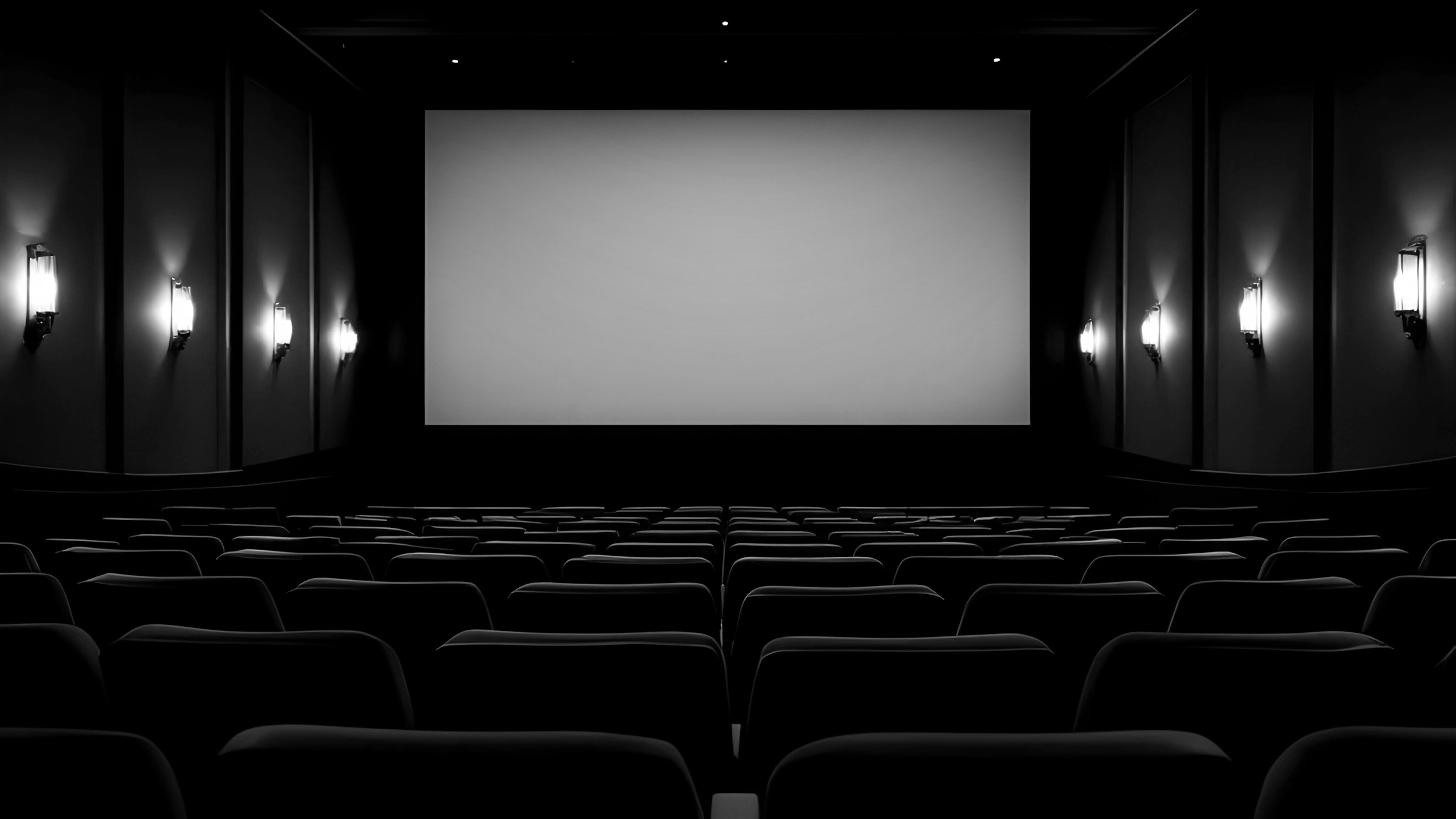 Empty movie theater with rows of seats facing a large blank screen, dim lighting, and wall-mounted lights along the walls.