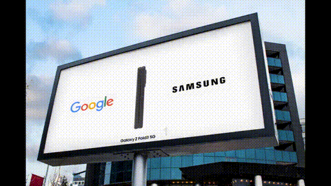 Large outdoor billboard showing Google and Samsung logos with a black vertical line in the middle, mounted on a building against a cloudy sky.