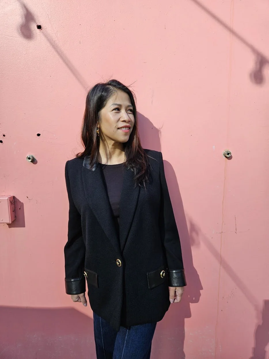 A woman with shoulder-length dark hair standing against a pink wall with holes and shadows, wearing a black blazer with leather cuffs and gold buttons.