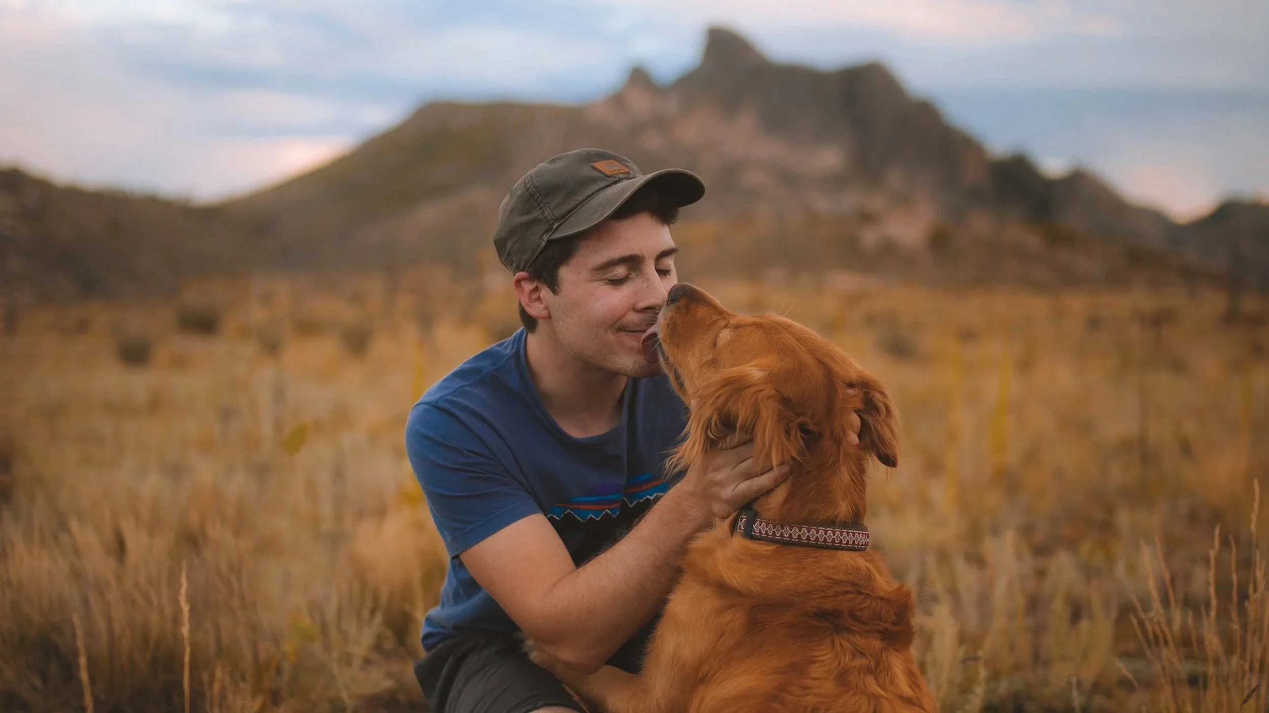 A man in a blue t-shirt and cap hugging a golden retriever in a field with mountains in the background.