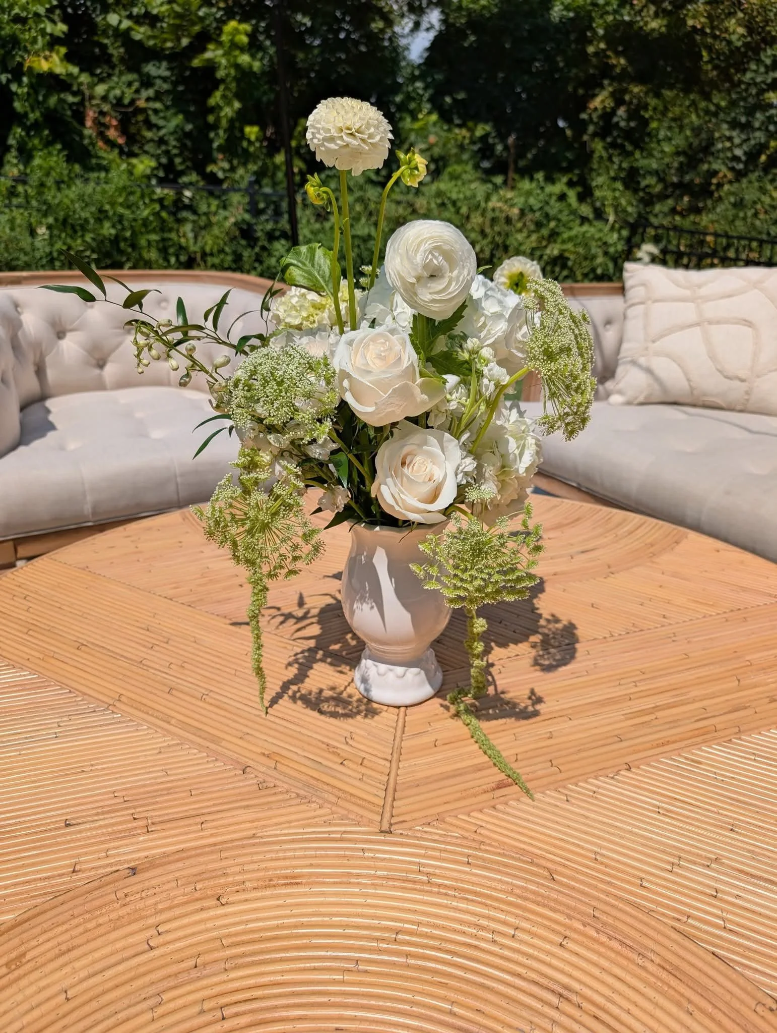 White flower bouquet in a white vase on a wooden table outdoors, with beige sofas and green trees in the background.
