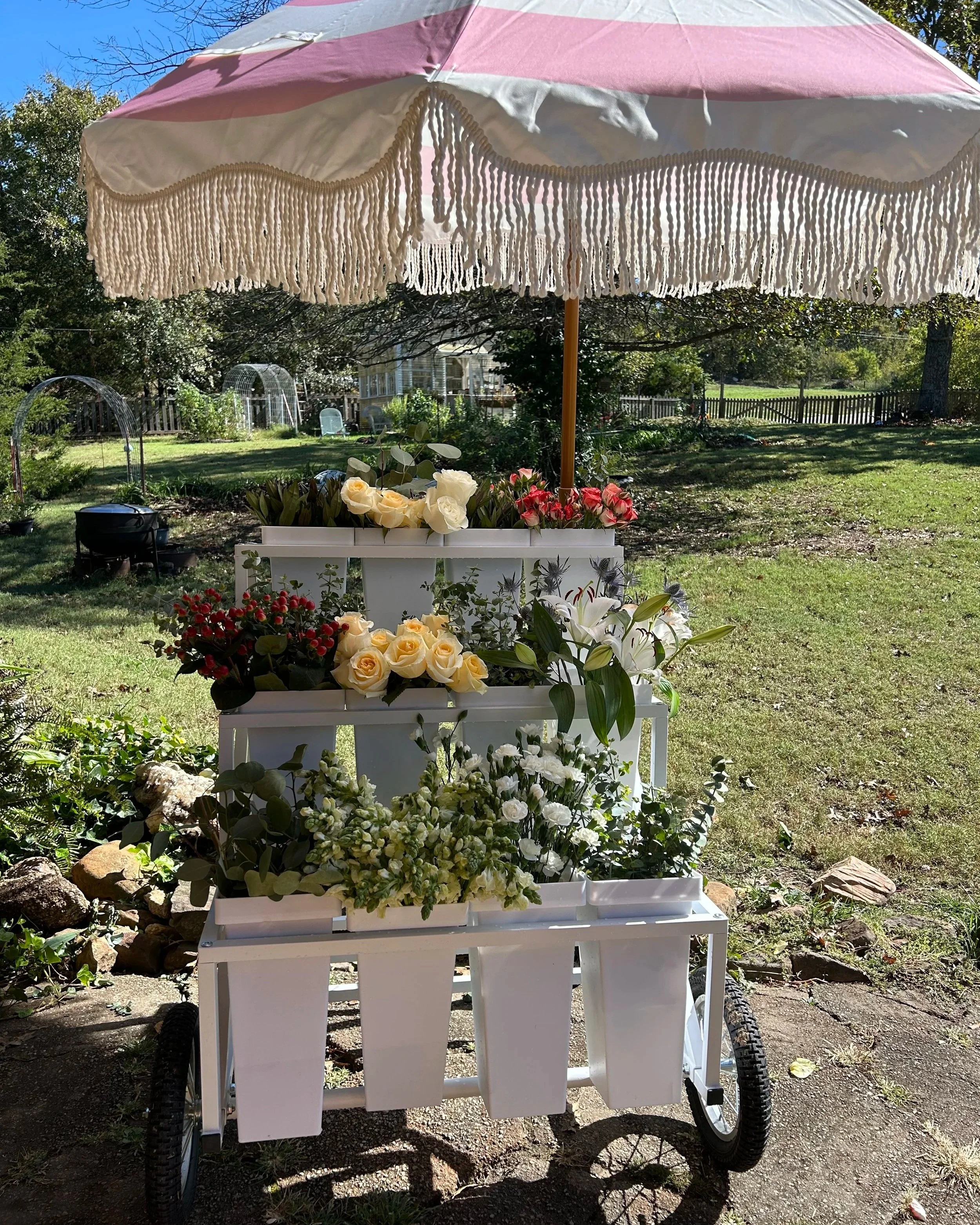 A white flower cart shaded by a pink and white parasol, filled with various flowers including roses, lilies, and other blooms, set in a garden yard with green grass, trees, and a fenced yard in the background.