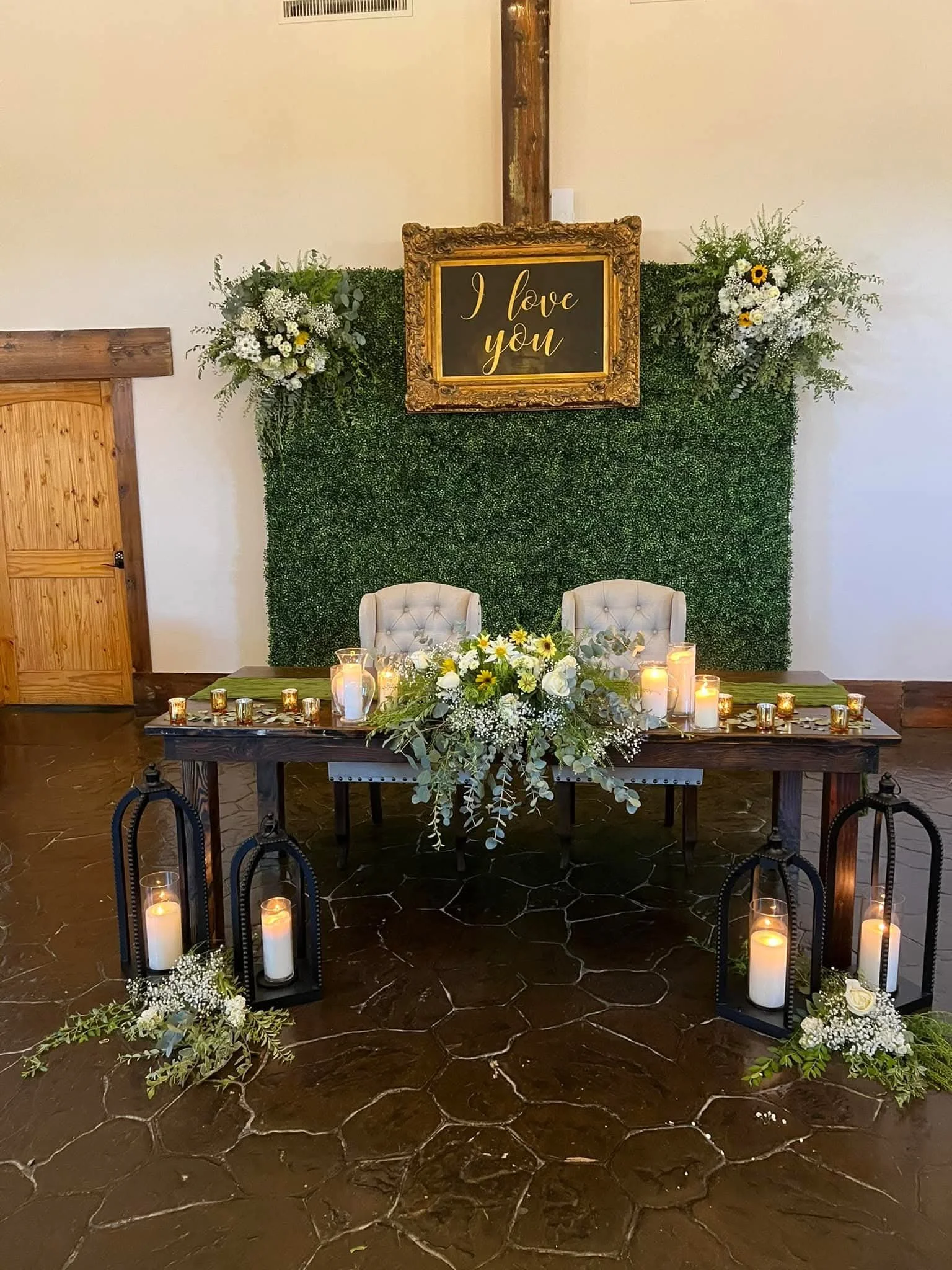 Decorated table with floral arrangement, candles, and framed sign reading 'I love you' on a green hedge backdrop with floral arrangements and candles on the floor.
