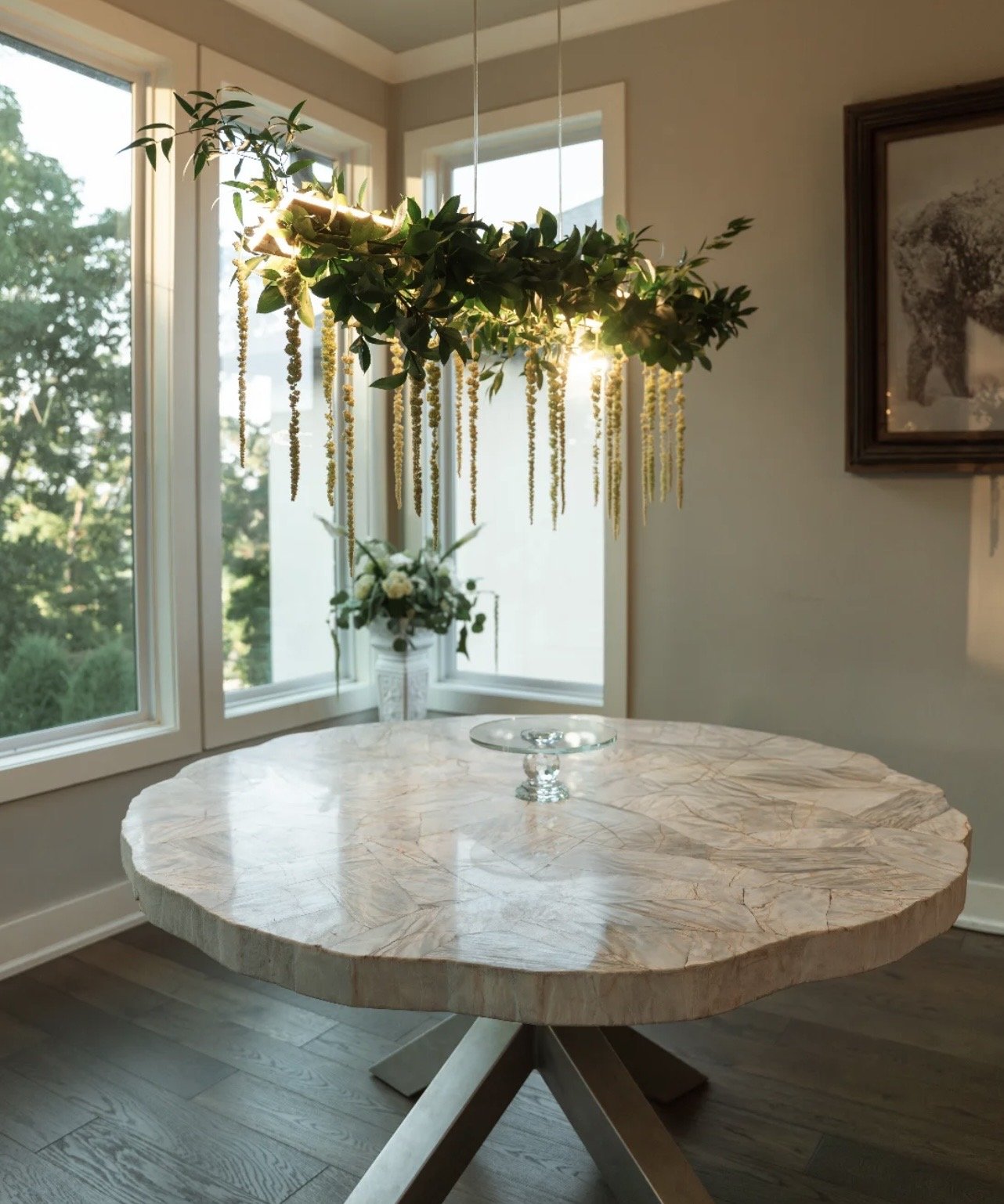 A round marble table with a glass cake stand, in a room with large windows and a hanging floral chandelier.