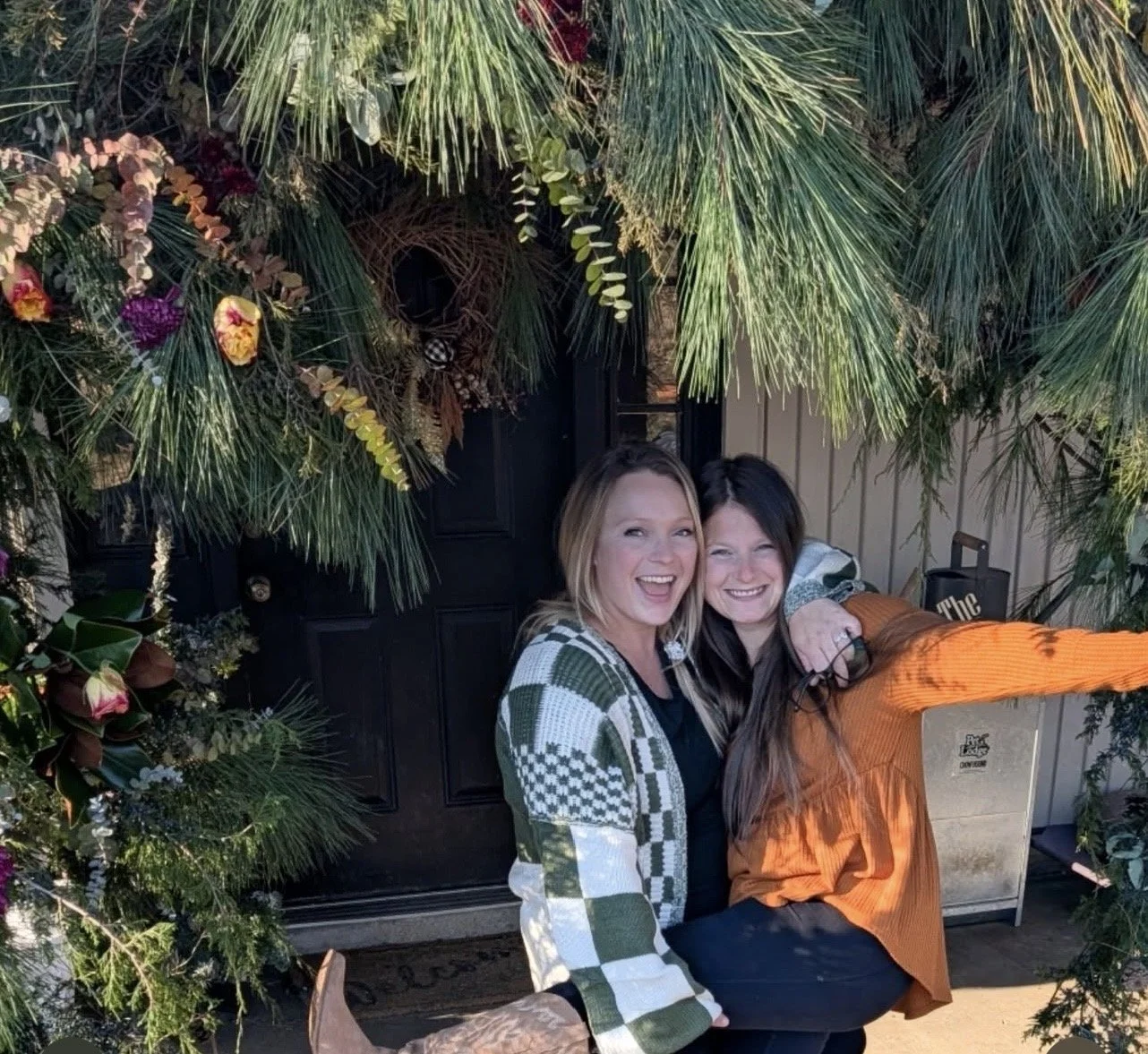 Two women smiling and taking a selfie in front of a decorated door with greenery and flowers.