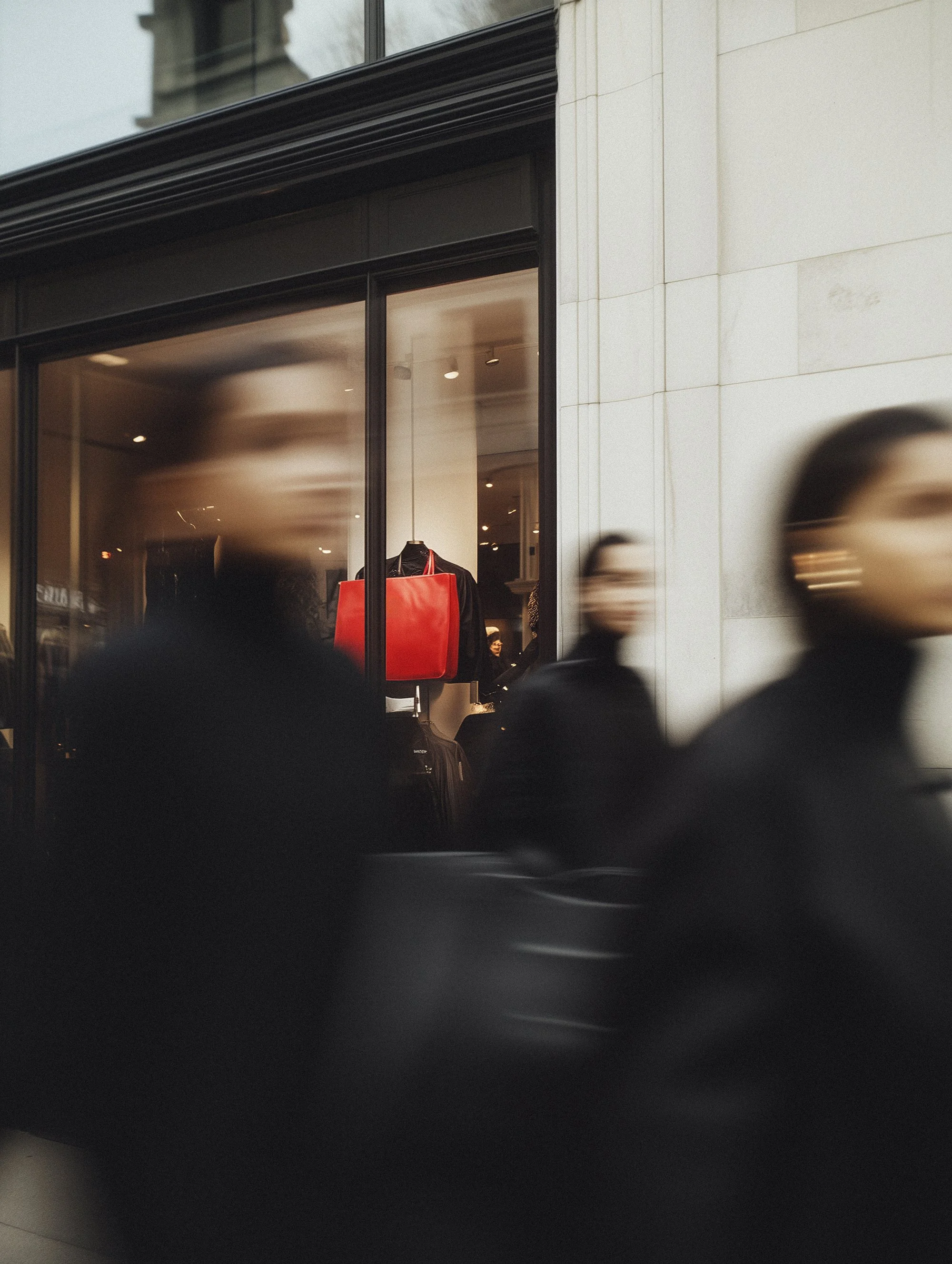 Blurry figures walking past a store window display featuring a red bag and black clothing.