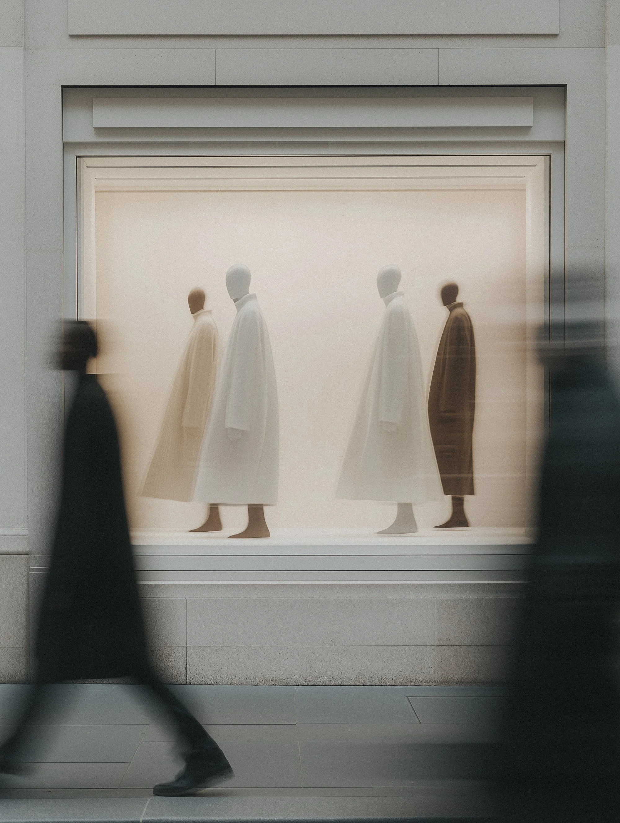 Blurred people walking past a shop window displaying mannequins in minimalist white and beige coats.