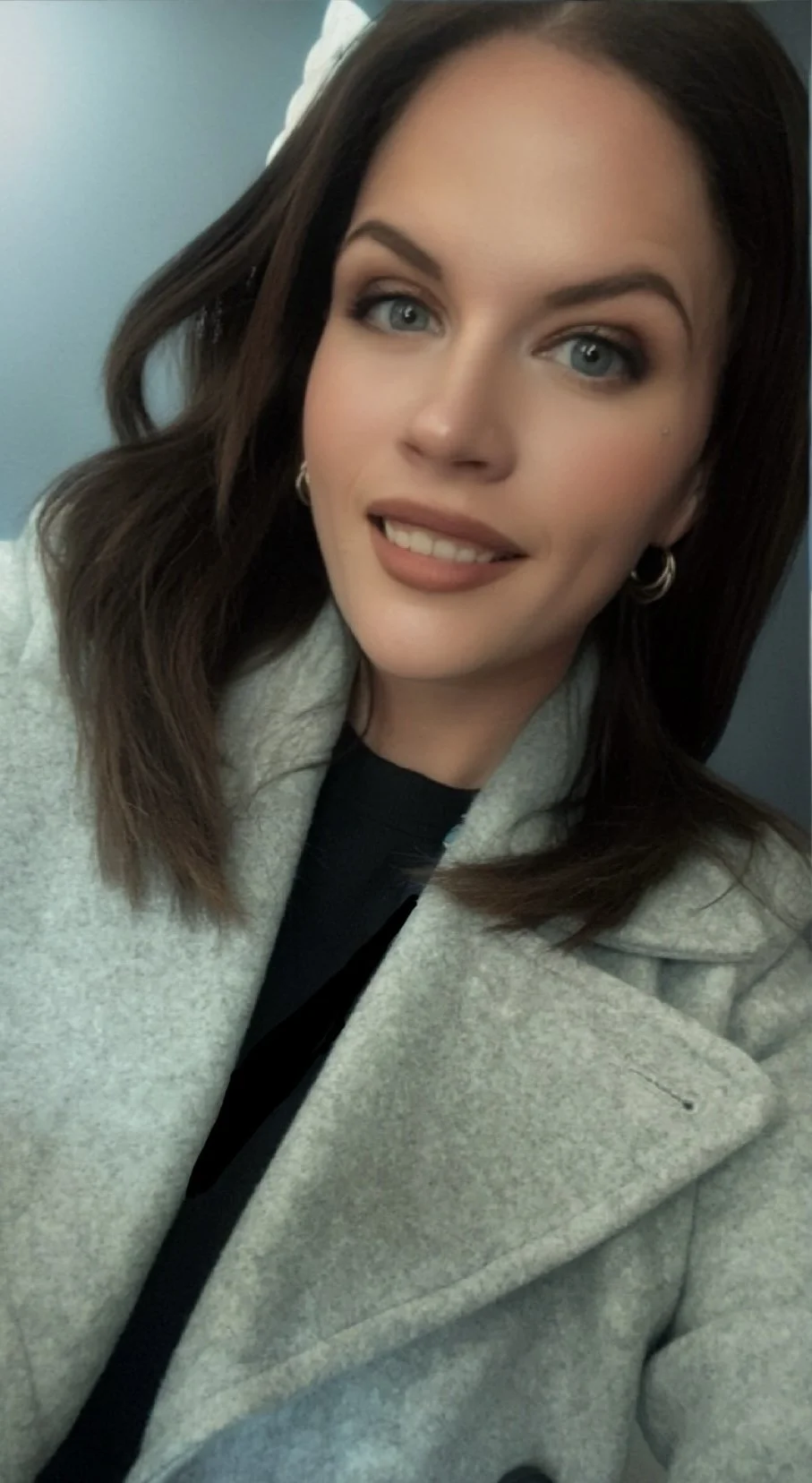 A young woman with long brown hair, blue eyes, and light makeup is smiling and looking at the camera. She is wearing a light gray coat over a dark shirt and has hoop earrings. The background shows a plain blue wall.