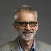 A middle-aged man with gray hair, a beard, and glasses, wearing a gray blazer and blue shirt, smiling against a plain gray background.
