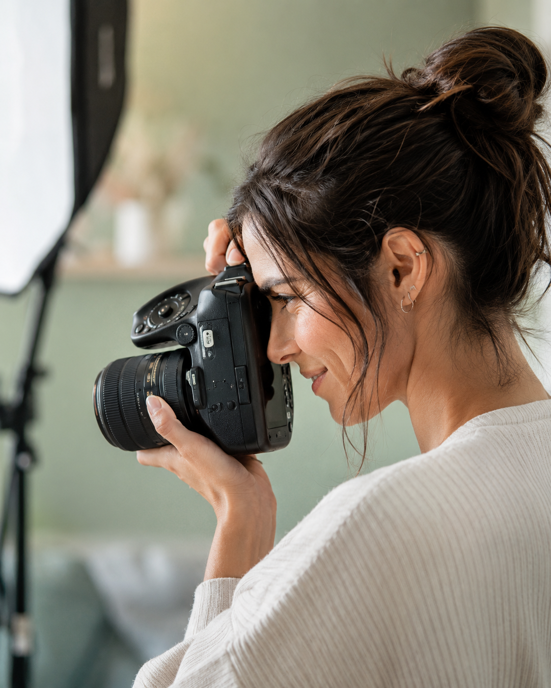 Une femme avec des cheveux bruns en chignon, tenant un appareil photo numérique et regardant à travers le viseur, en studio avec un fond vert.