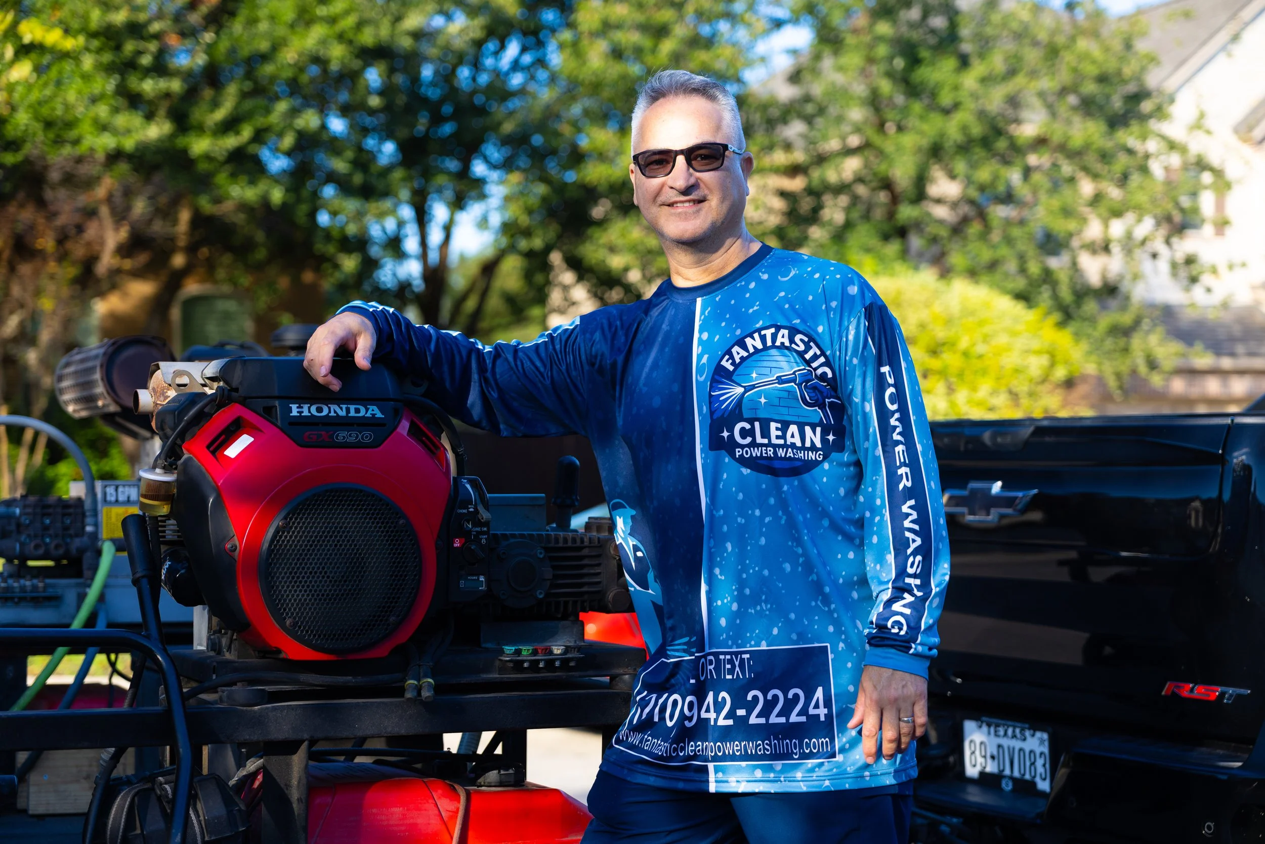 A man in sunglasses wearing a blue shirt with white text and graphics, standing outdoors next to a piece of equipment with the brand Honda. The background includes trees and a black pickup truck.