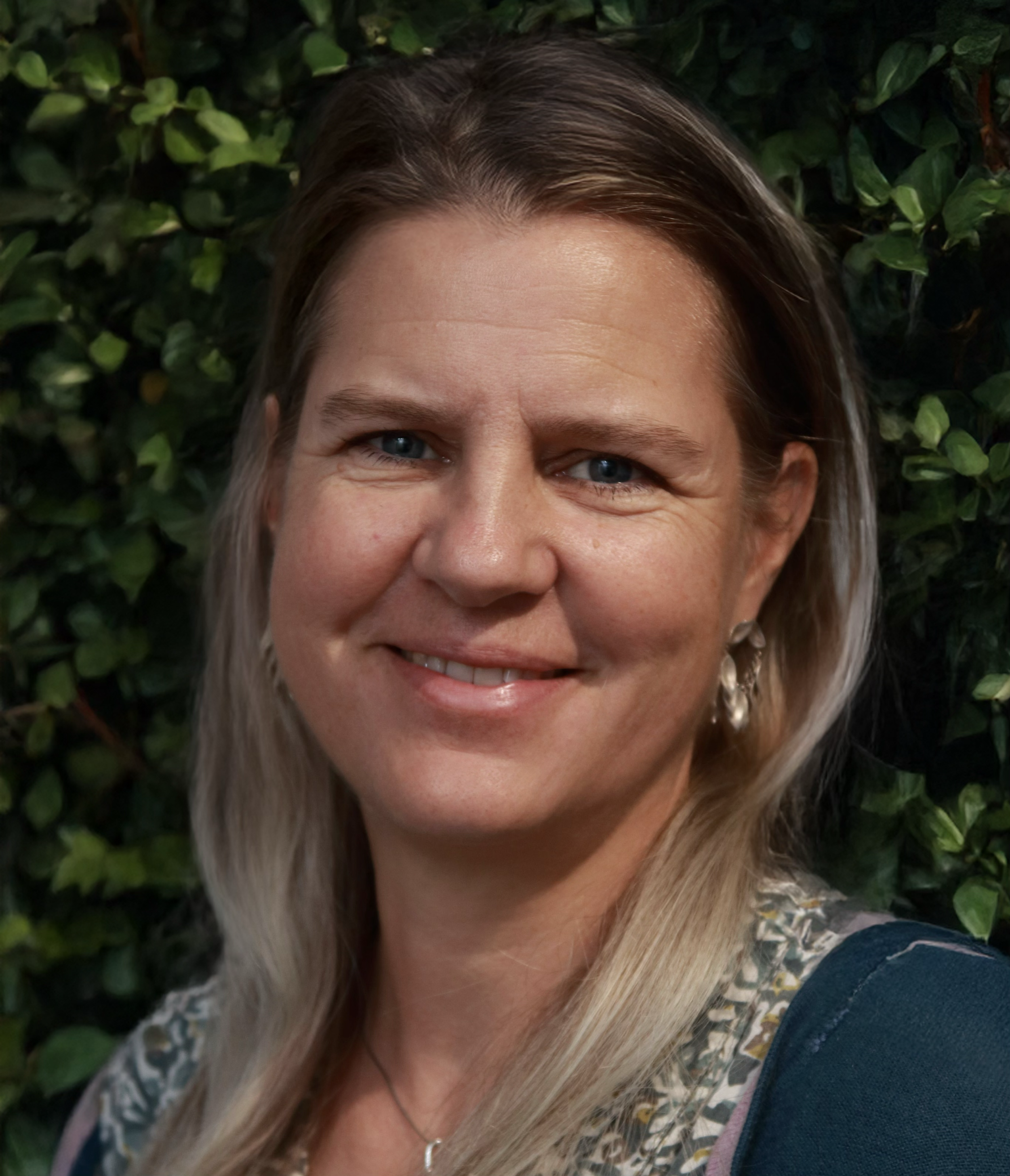 Portrait of a smiling woman with blue eyes and blonde hair, standing outdoors in front of green foliage.