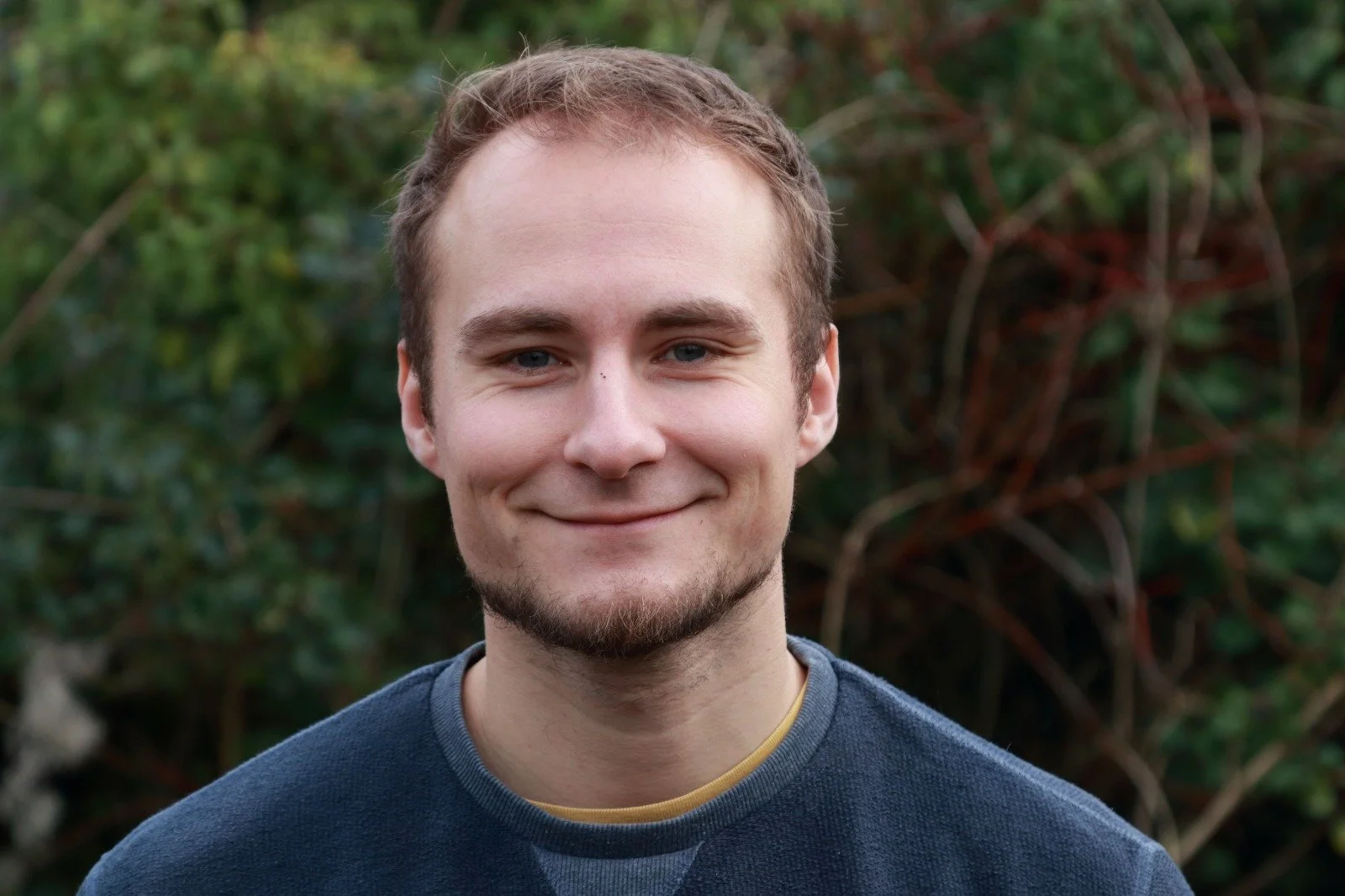A young man with light skin, short light brown hair, blue eyes, and a light beard, smiling outdoors with greenery in the background.