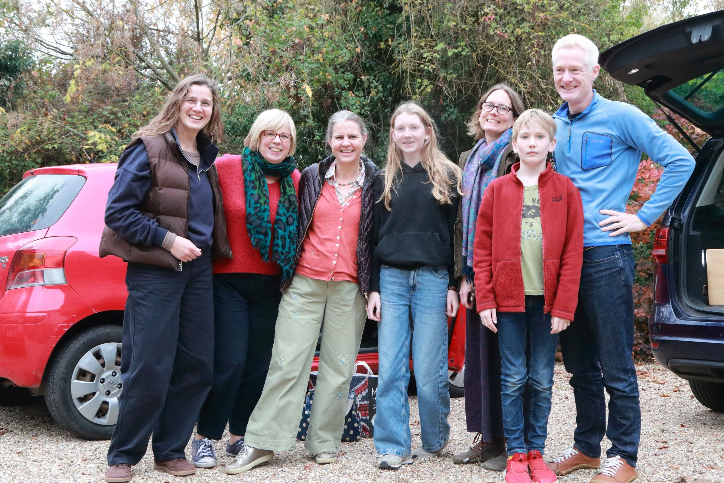 A group of eight people standing outdoors in front of a wooded background, smiling for the photo. Two cars are visible, one red on the left and one black on the right, with open trunk.