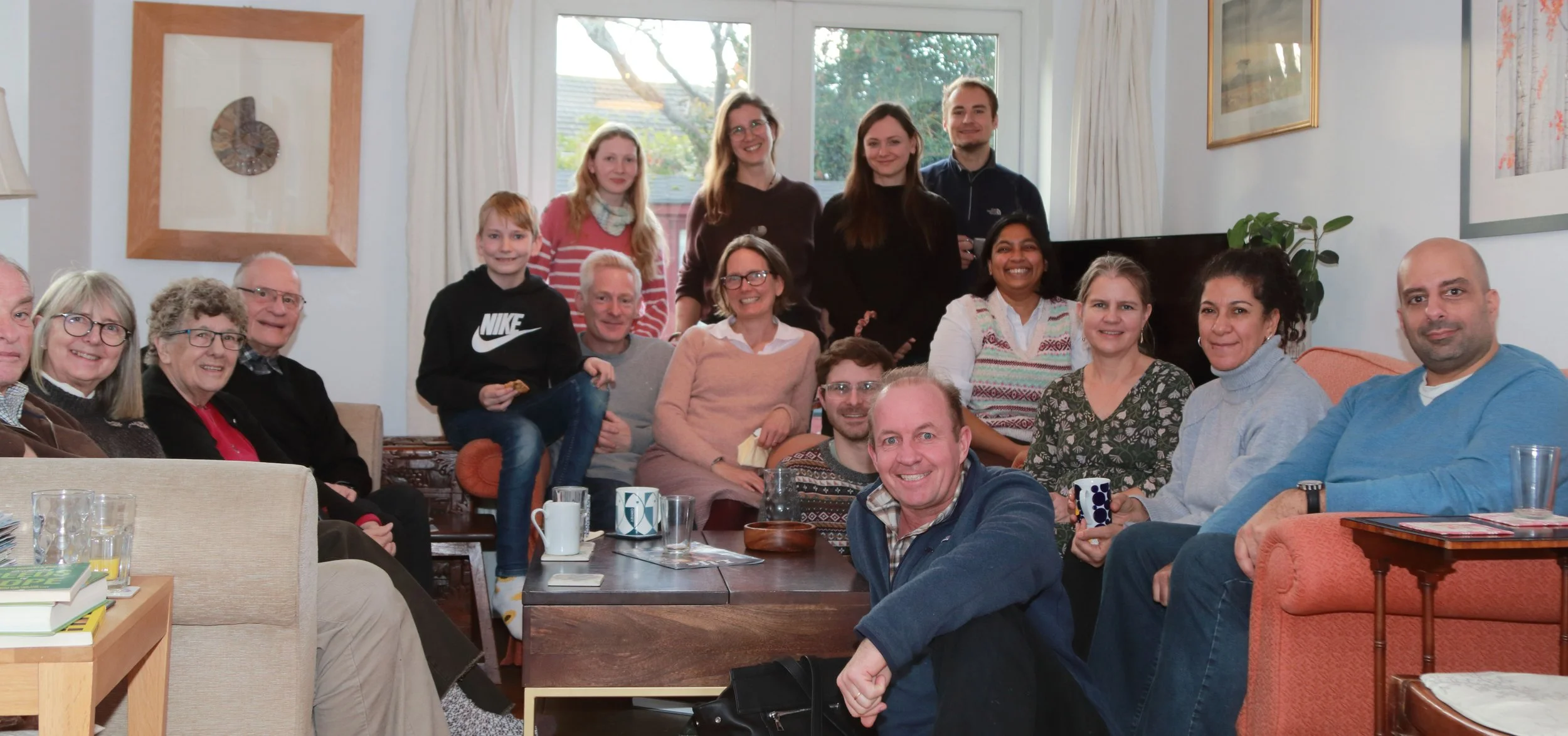 A group of 16 people gathered in a living room for a group photo, with some seated on sofas and others standing behind. They are smiling and appear to be enjoying each other's company, with a large window and artwork visible in the background.