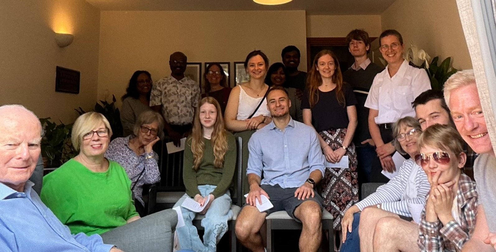 Group of people gathered indoors, smiling, with some sitting and others standing behind them, of assorted ages.