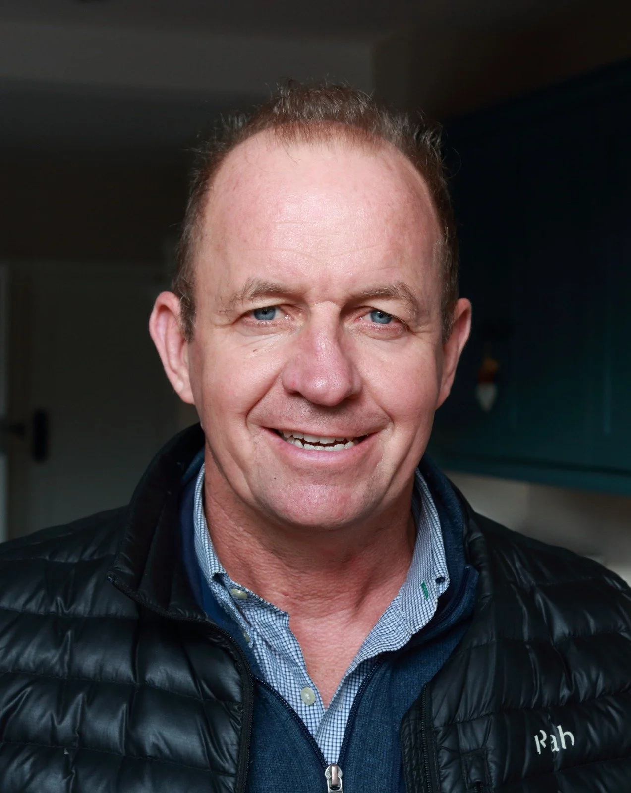 Close-up portrait of a smiling middle-aged man with short hair, wearing a black quilted jacket and a blue collared shirt.