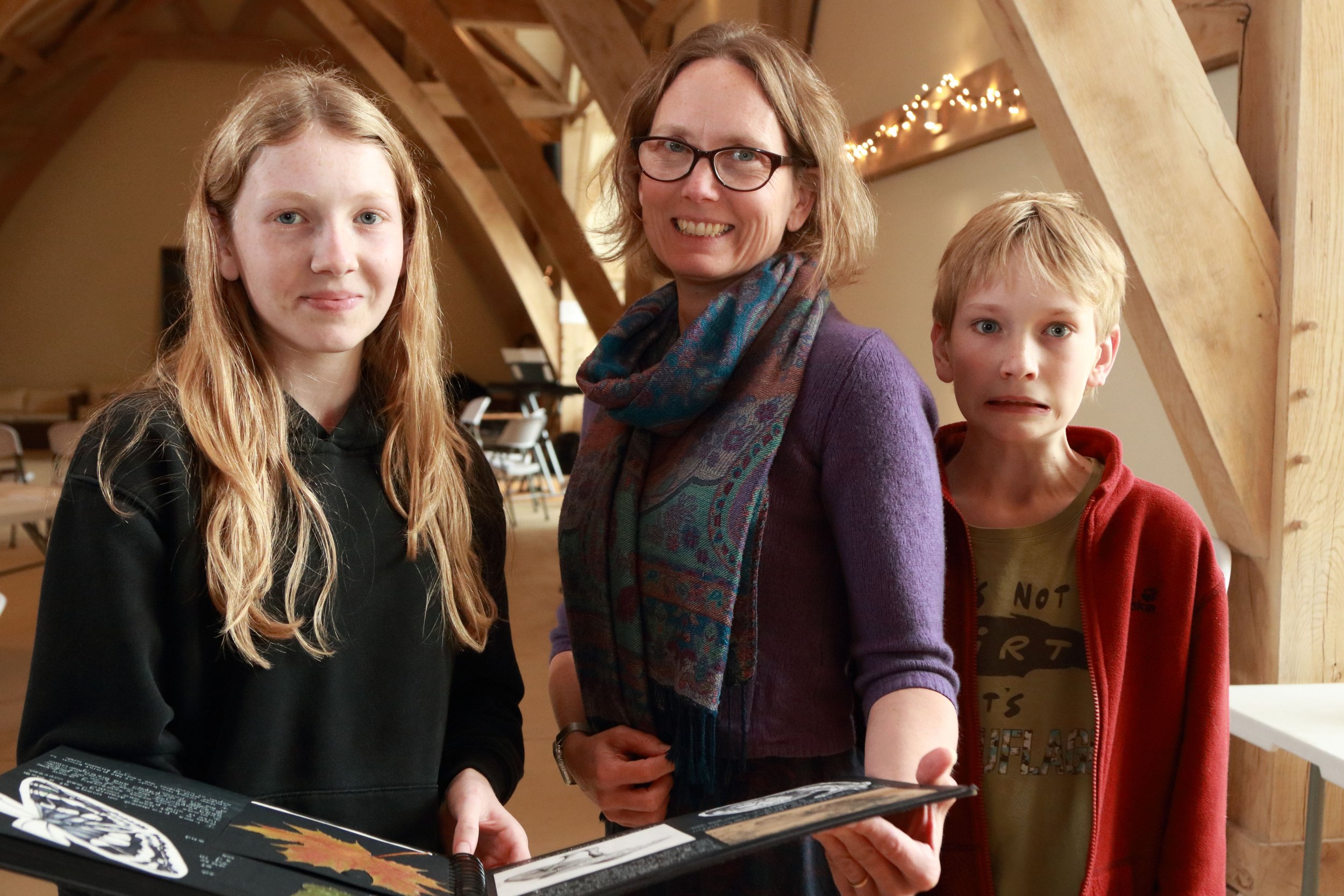 A woman with glasses and two children, a girl and a boy, standing indoors with wooden beams and string lights in the background. The woman is smiling, the girl has a neutral expression, and the boy appears surprised.
