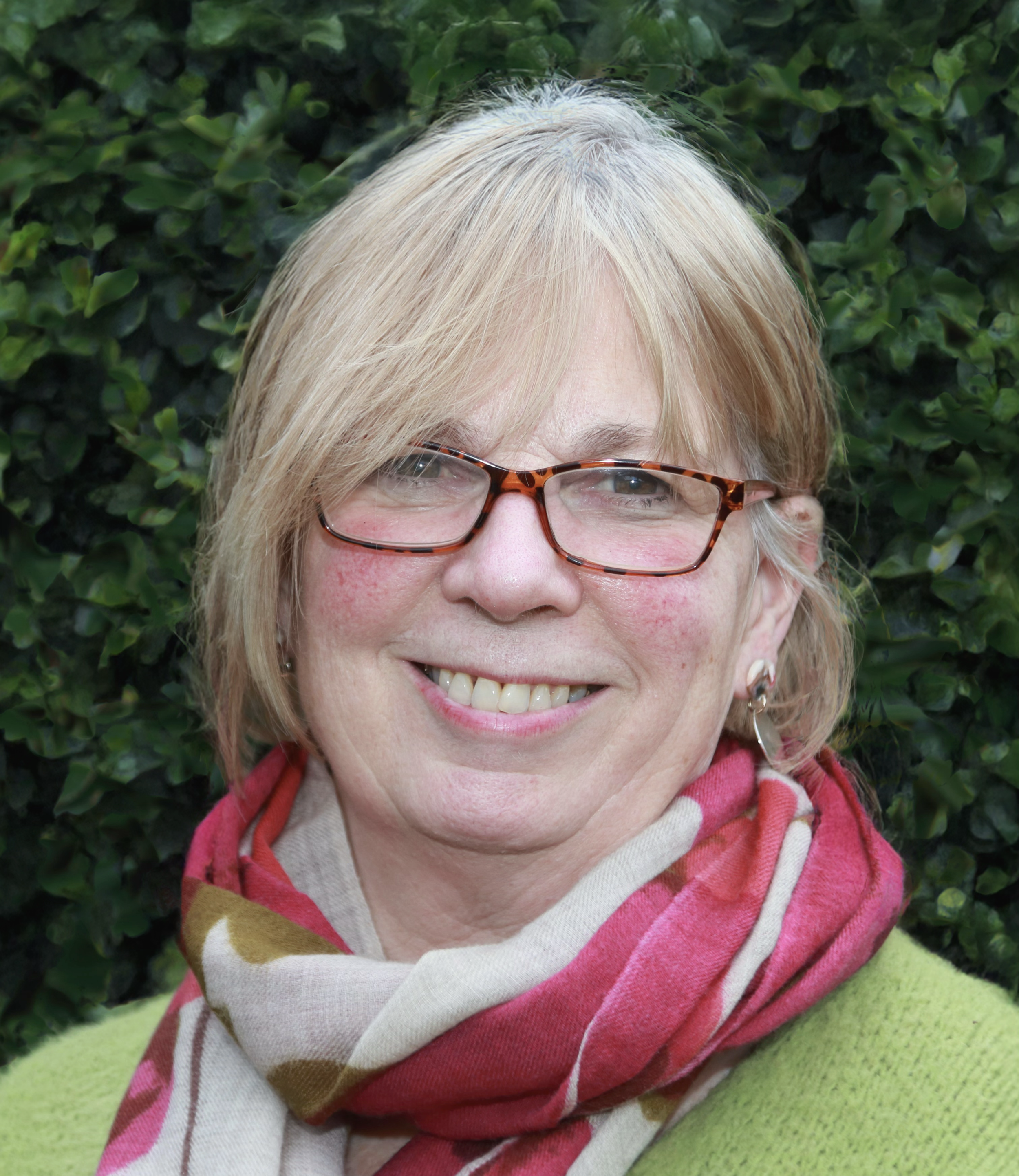 A middle-aged woman with blonde hair, glasses, and earrings smiling outdoors with a leafy green background.