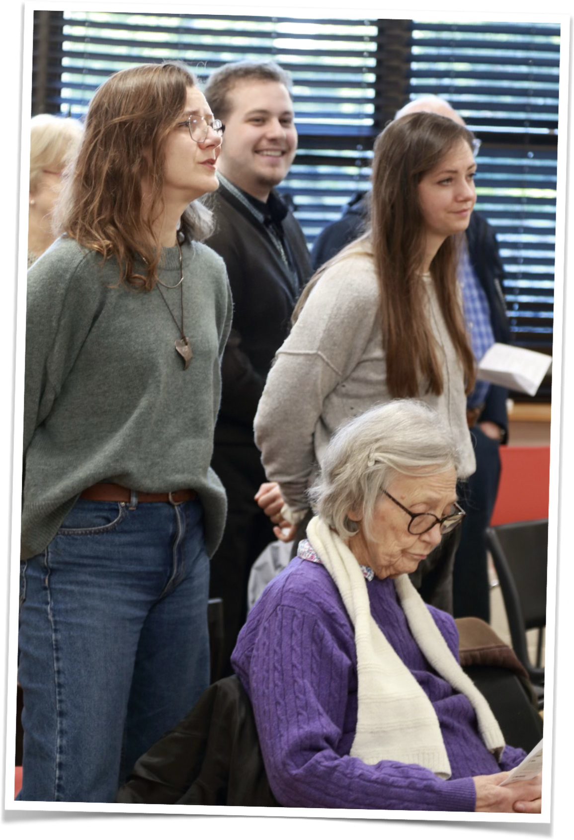 A group of people attending a church service, with some standing and one older woman sitting, looking at the song sheet.