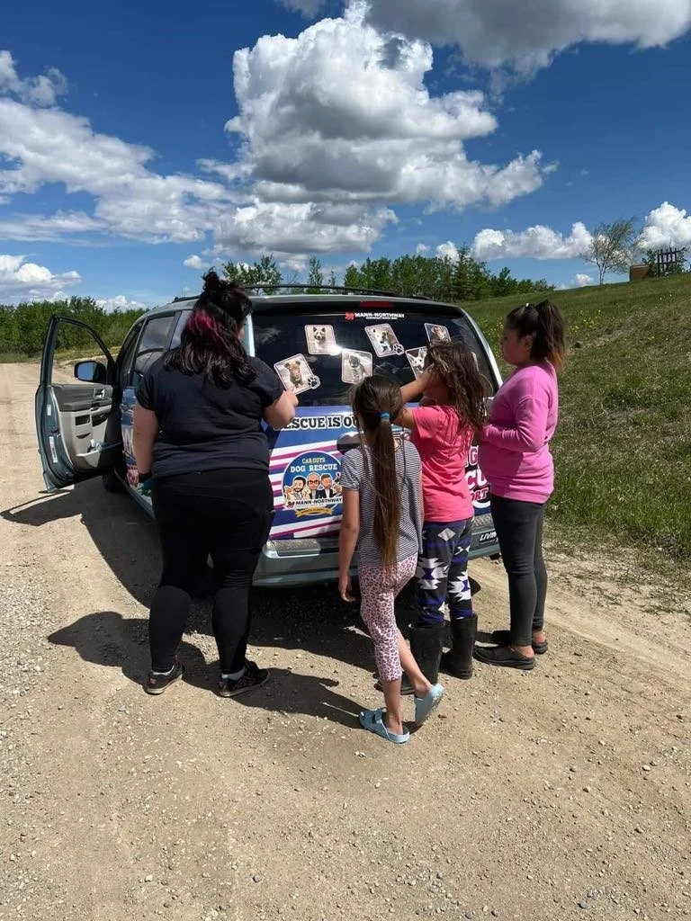 Group of children and woman gathered around a dog rescue van outdoors on a dirt road, with a partly cloudy sky and green grass in the background.