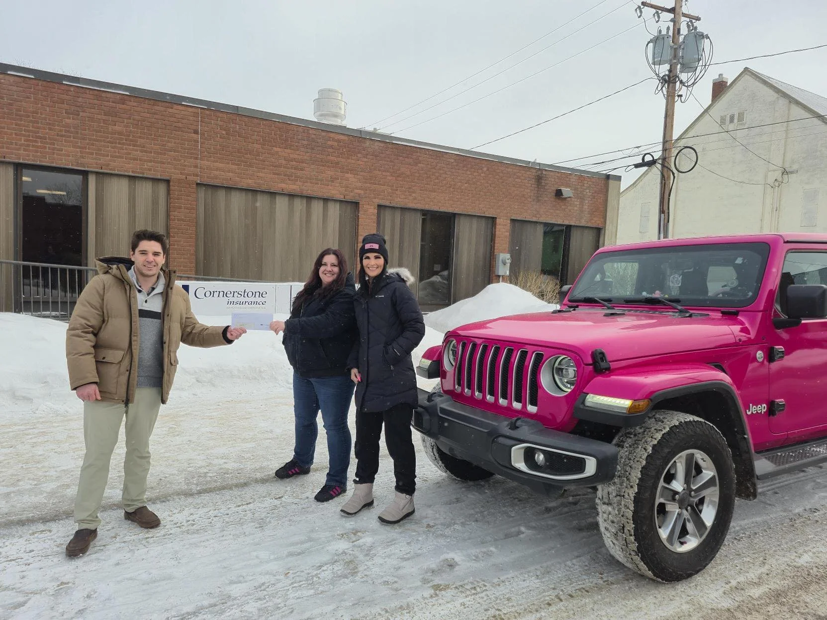 Three people standing outdoors in winter, holding a check, next to a bright pink Jeep SUV on a snowy street, with a brick building and utility pole in the background.