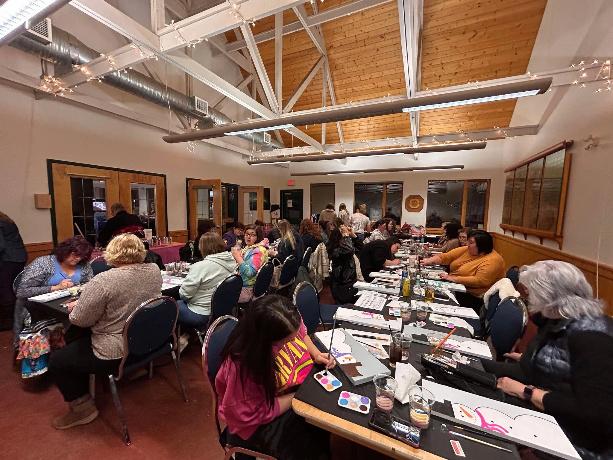 People seated at long tables painting and socializing in a well-lit room with wooden ceiling and paneled walls.