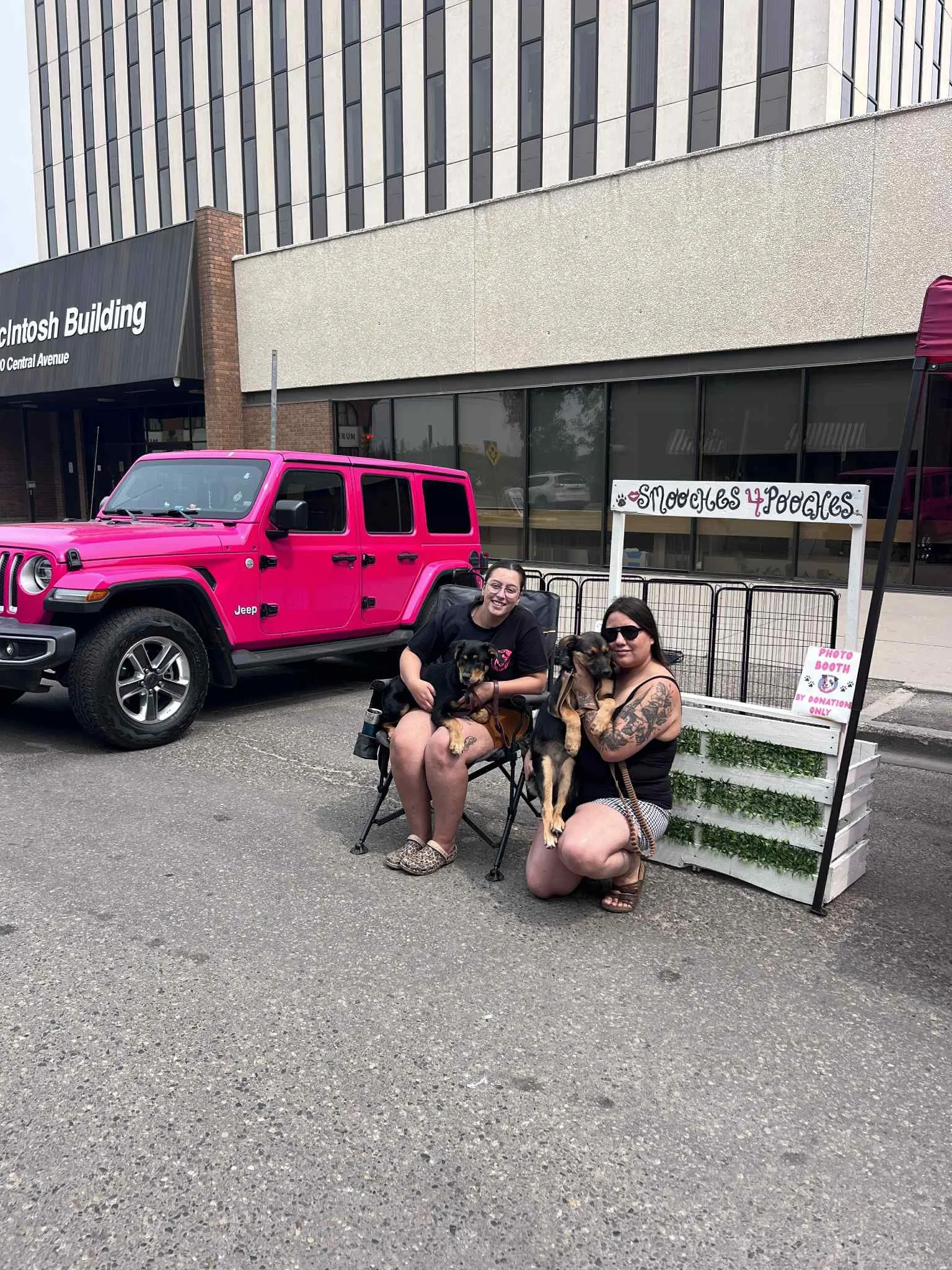 Two women sitting on chairs outside a building with a sign next to them that reads 'Snuggles & Poodles'. They are holding large dogs, and there is a bright pink Jeep parked nearby.