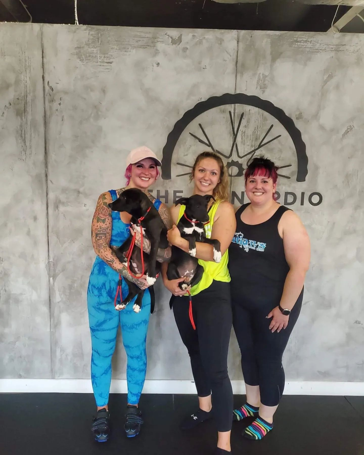 Three women smiling and holding young black puppies with white markings. They are standing in front of a gray wall with a clock and the text 'THE SOUND RADIO'.