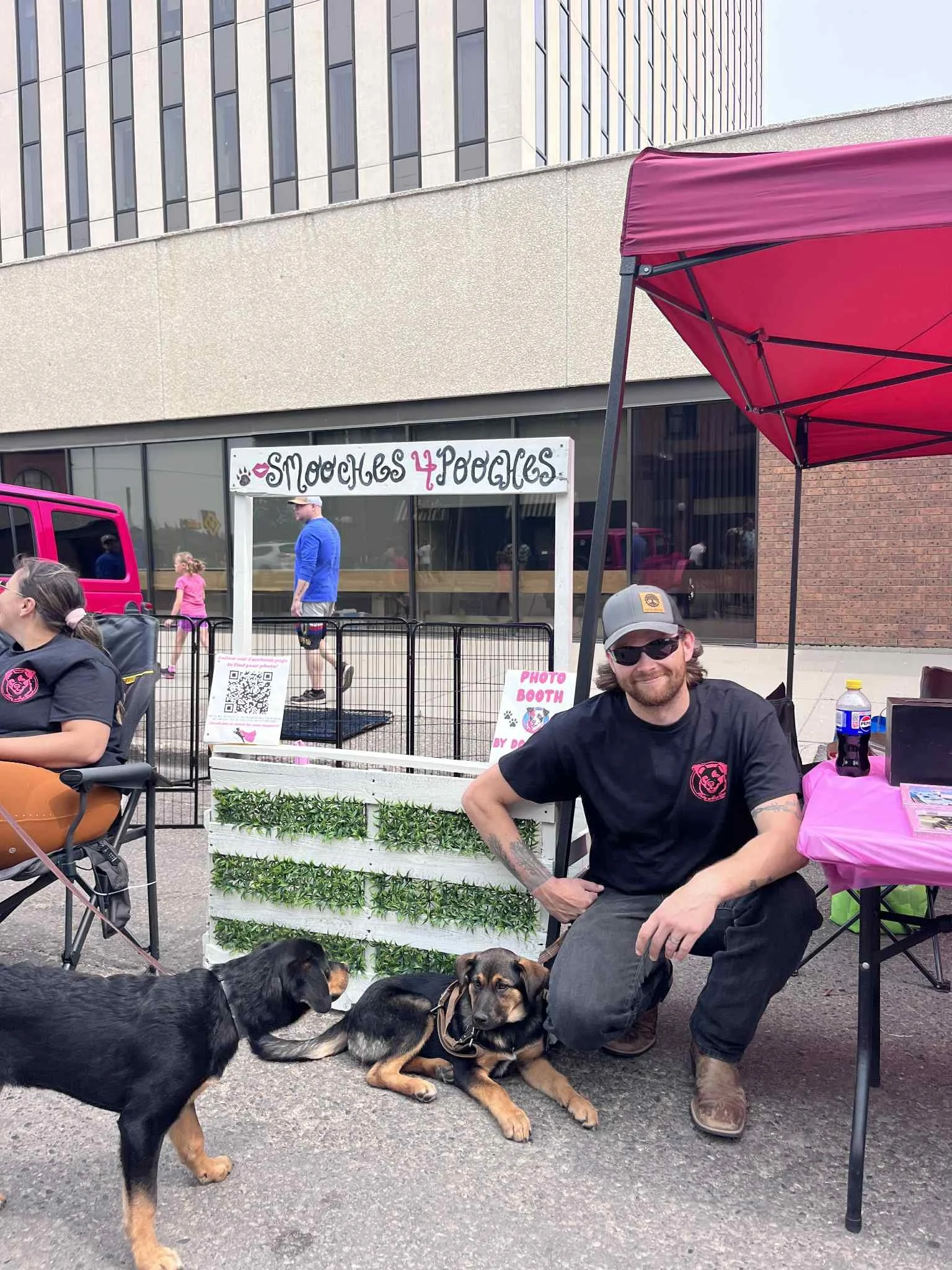 Man with sunglasses kneeling with two dogs at a booth called 'Moches 4 Pooches' at an outdoor event.