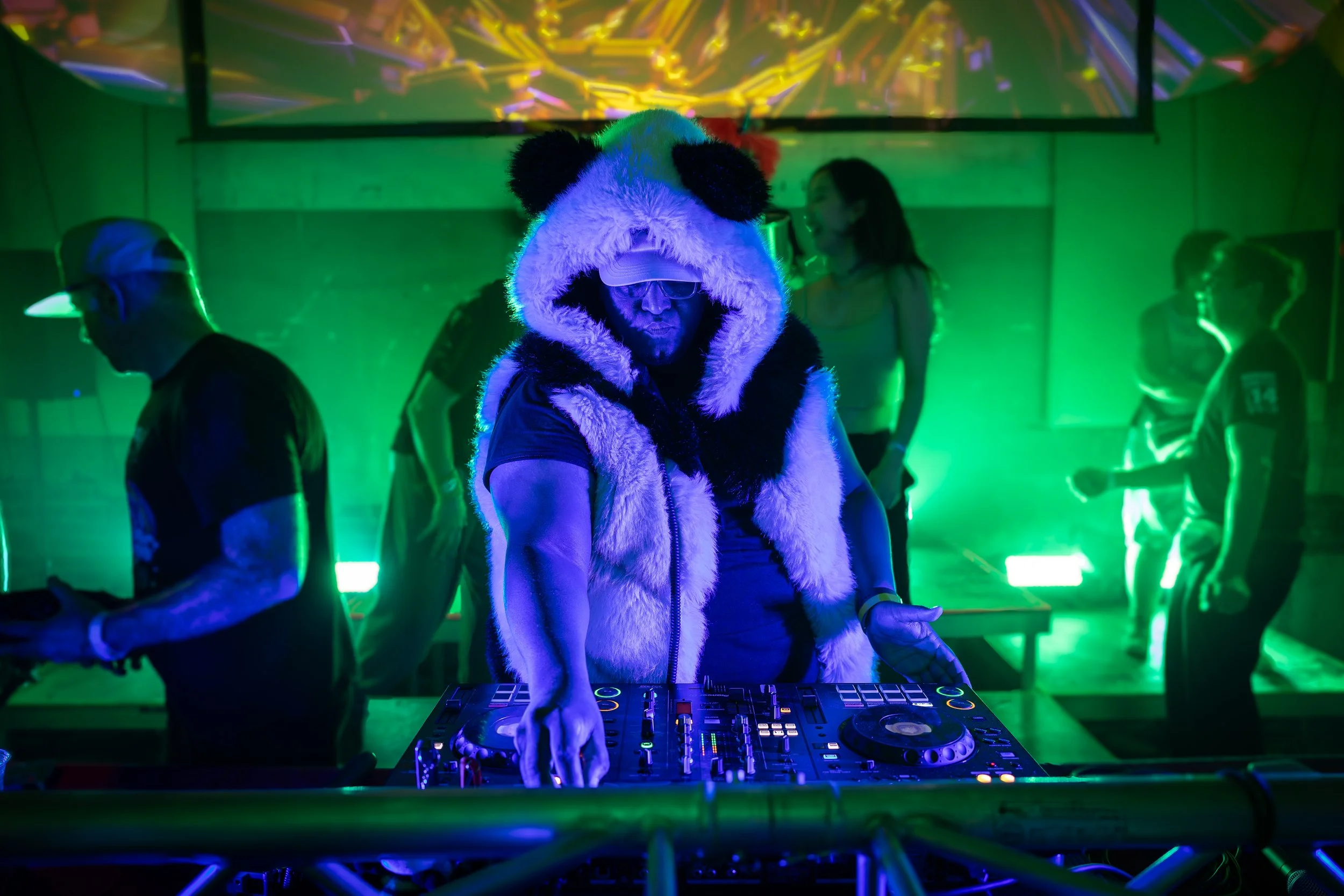 Person wearing a panda costume hooded jacket DJing at a nightclub with green lighting and several people dancing in the background.