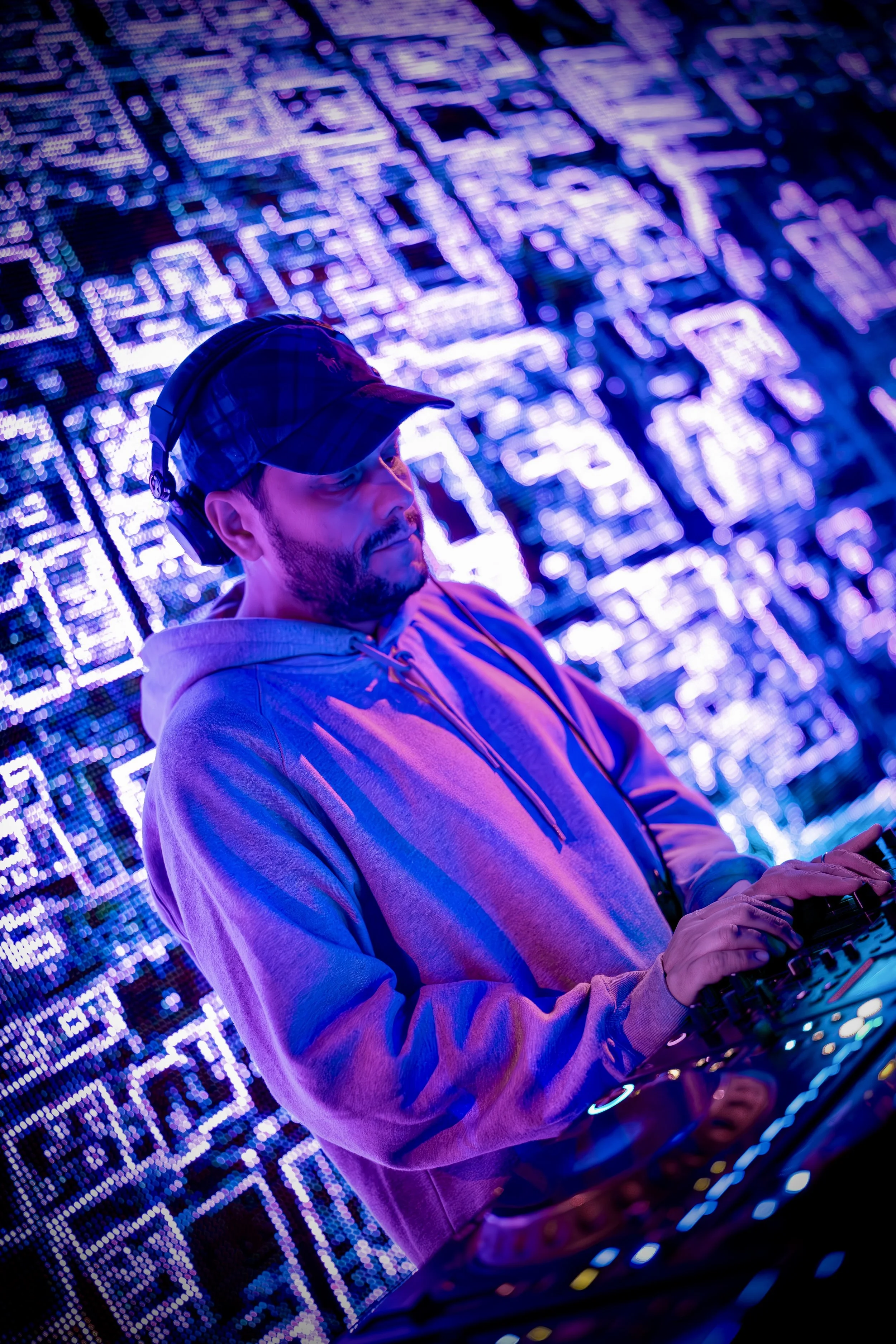 A DJ wearing a hoodie, baseball cap, and headphones, performing with electronic music equipment in front of a backdrop of colorful, neon LED lights.