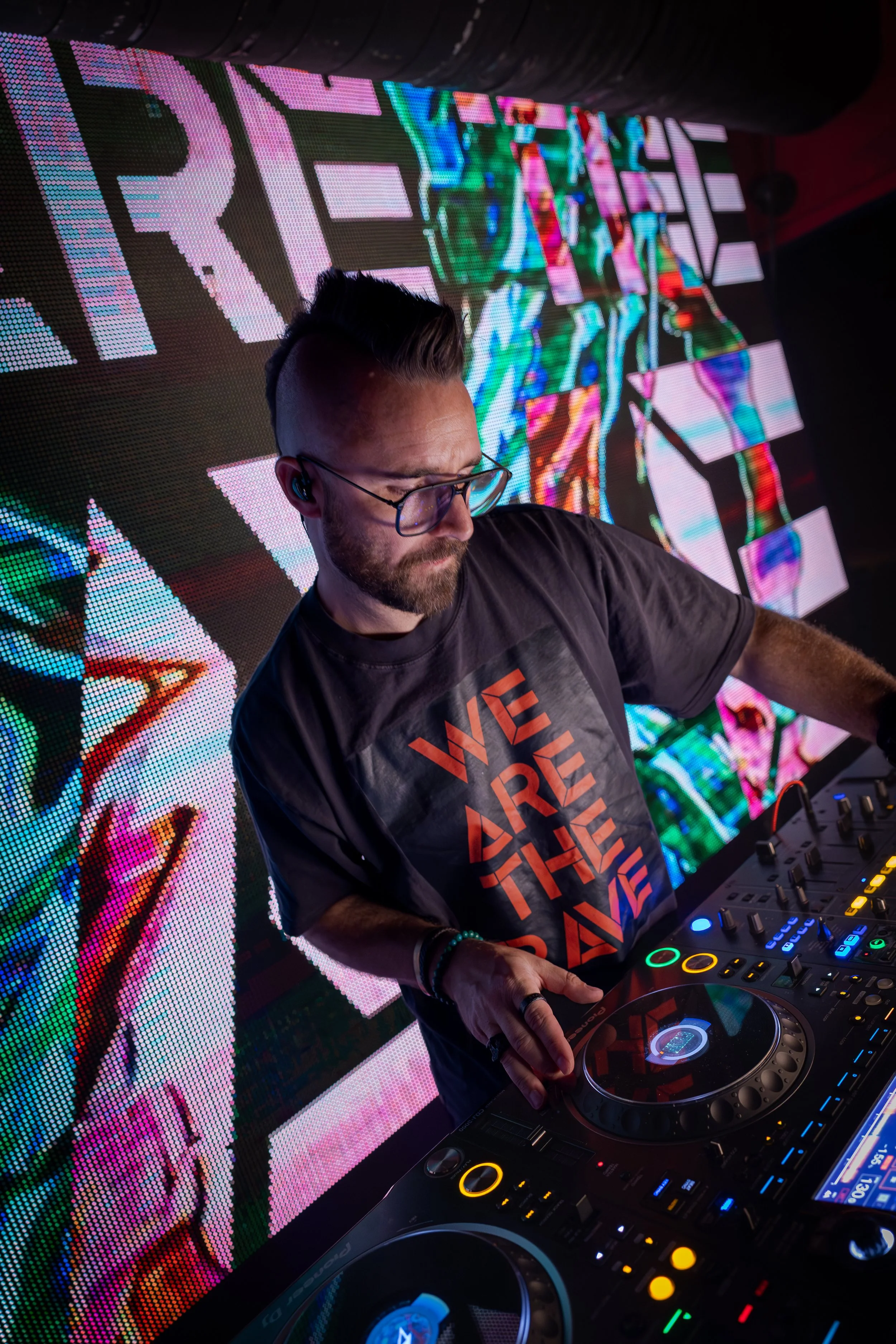 DJ wearing glasses and a dark T-shirt with the text 'WE ARE THE RAVE,' DJing with colorful LED screens displaying vibrant abstract graphics in the background.
