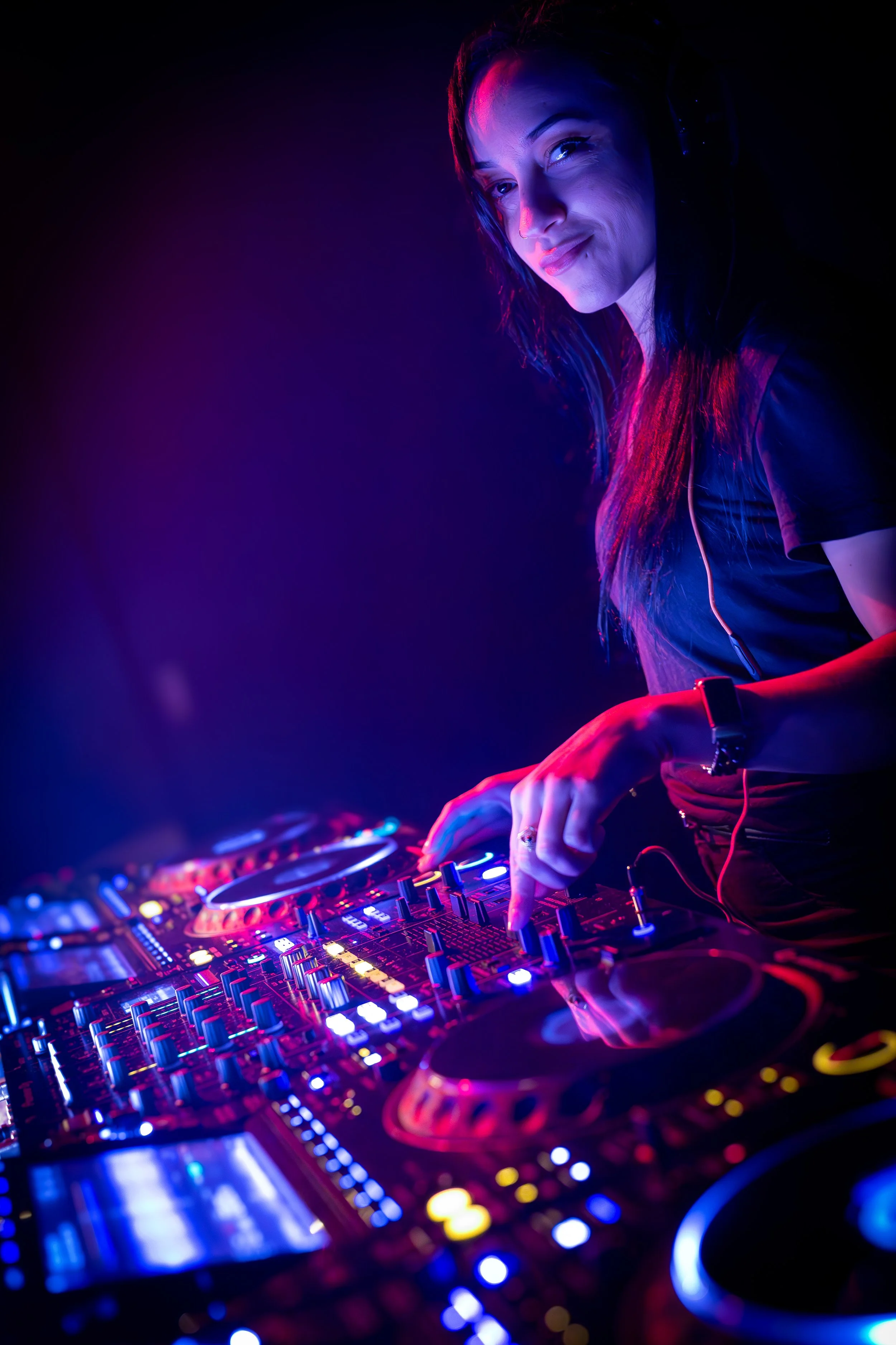 A female DJ wearing headphones, standing behind a DJ mixer with colorful lights, smiling at the camera.