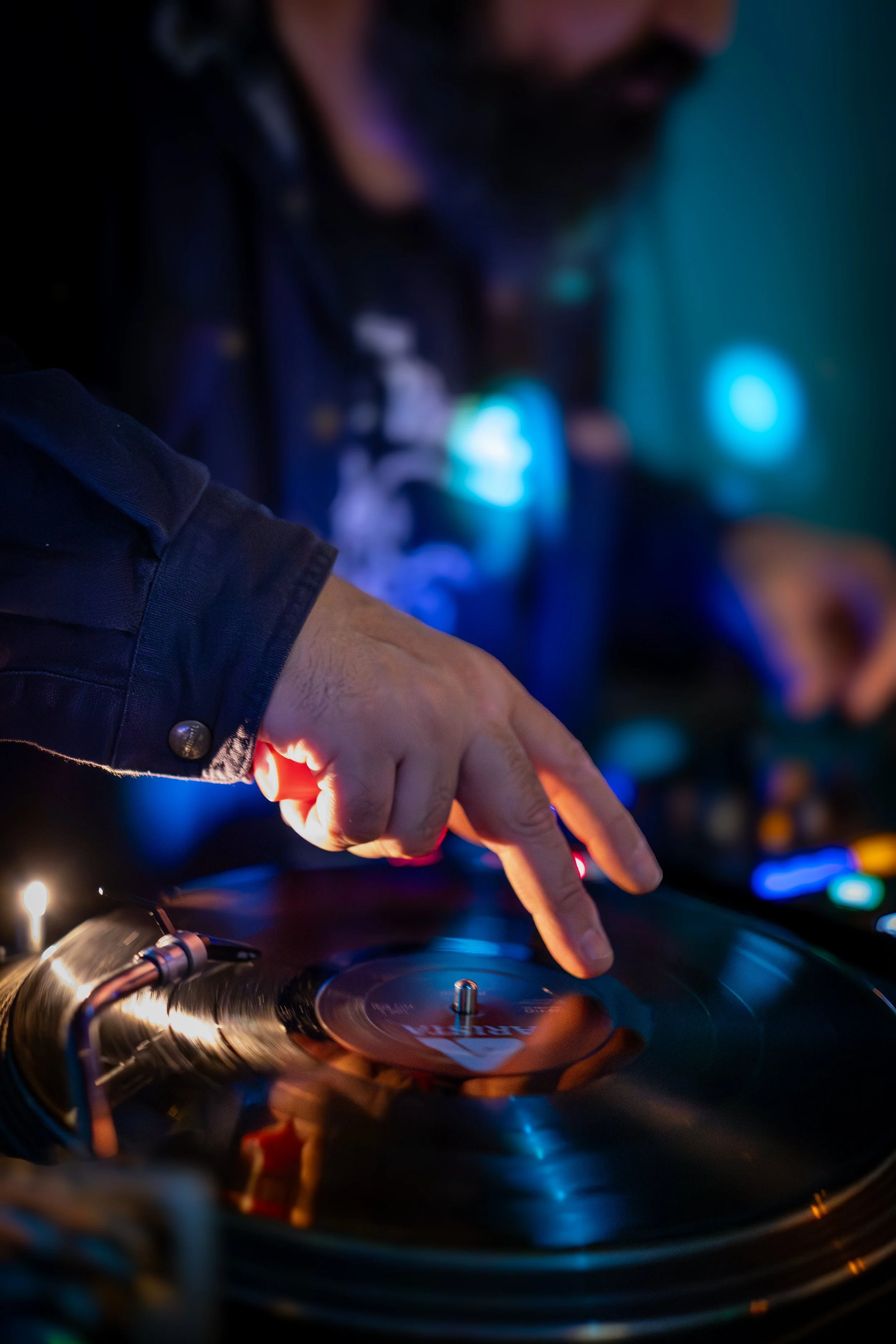 Close-up of a person DJing, with their hand on a vinyl record on a turntable, illuminated by colorful club lights.