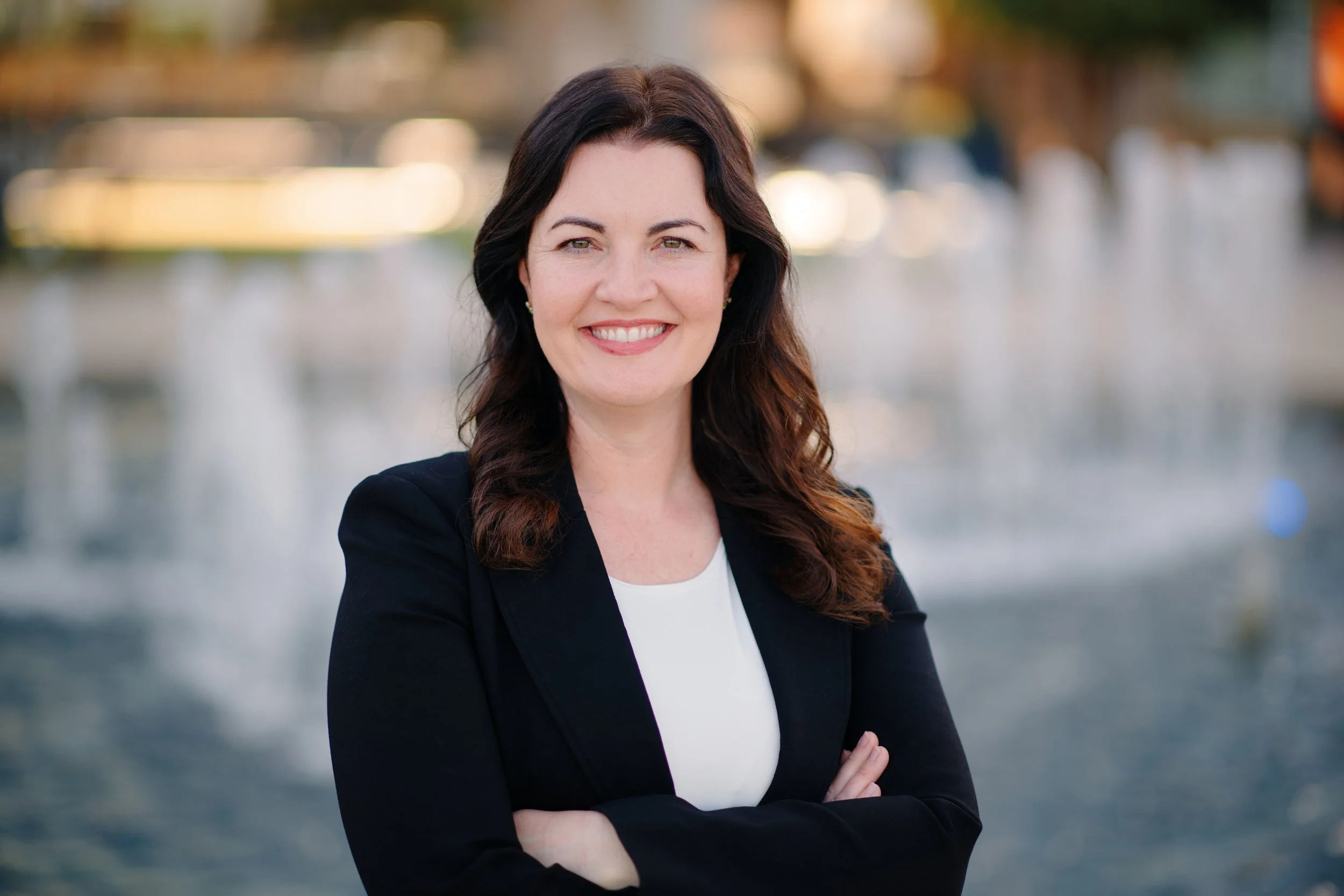 Katie O'Brien Duzan, a woman with dark wavy hair, wearing a black blazer and white shirt smiling with arms crossed outdoors in front of Show Fountain in the Entertainment District of Arlington, Texas.
