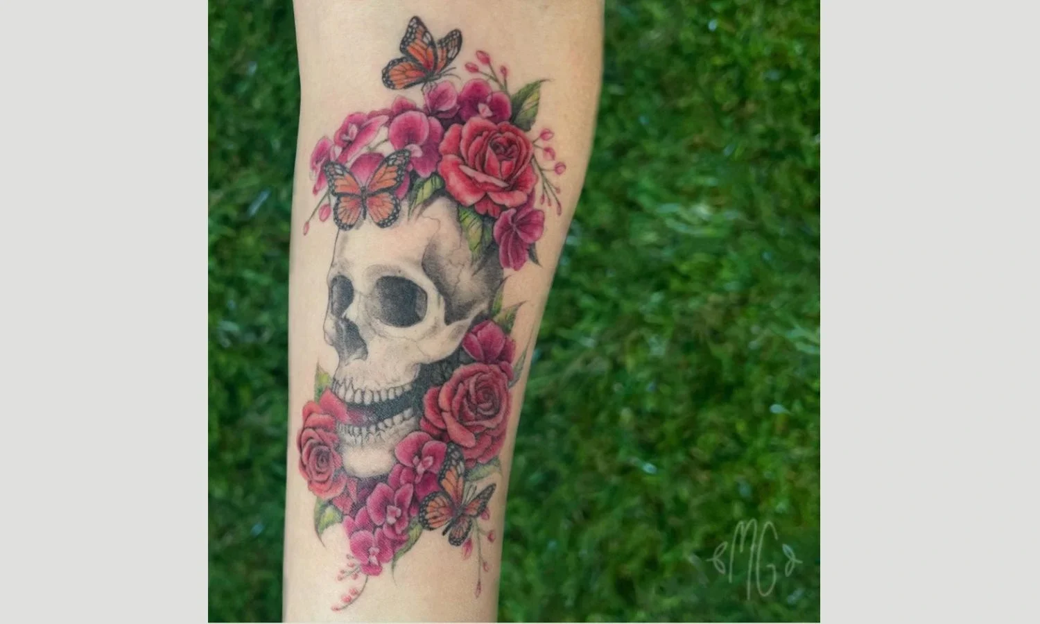 Colorful skull tattoo surrounded by red roses, pink flowers, green leaves, and butterflies on the forearm.