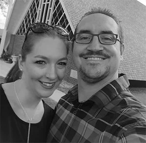 A smiling man and woman taking a selfie outdoors, with a building in the background.