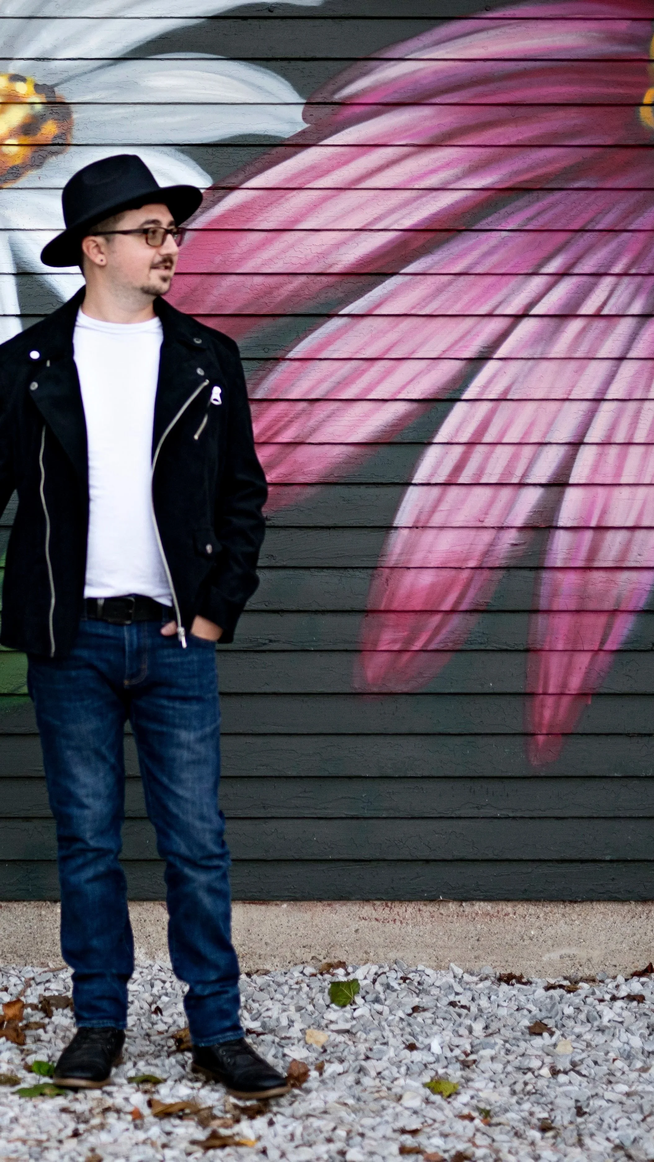 A man in a black jacket and fedora stands in front of a mural of pink and white flower petals painted on dark wooden planks.