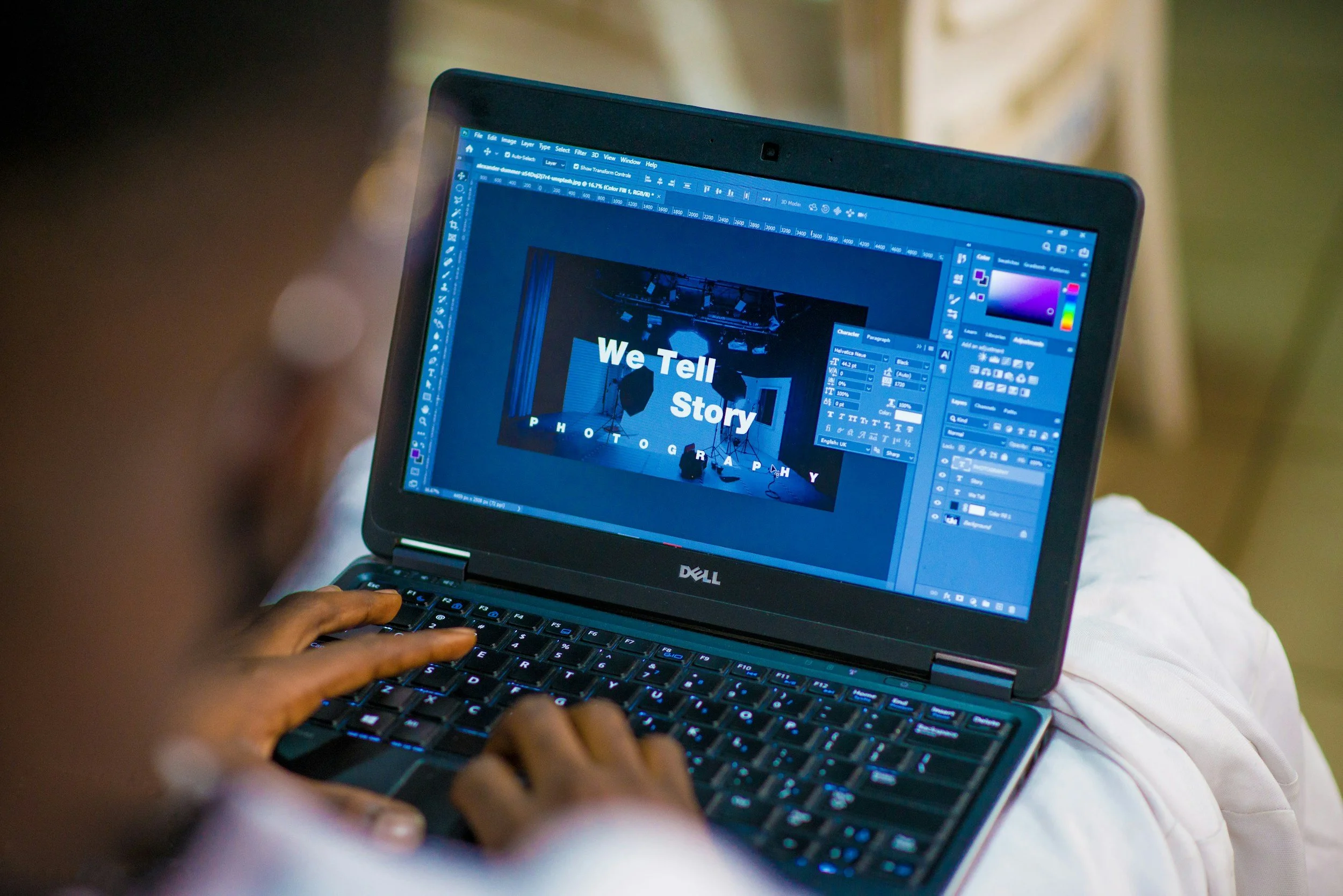 A laptop screen displays a graphic design project with the text "We Tell Story" and "Photography" visible, with tools and layers of graphic design software on the screen, and a person using the keyboard.