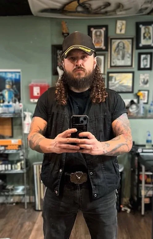 A man with curly hair, tattoos, and a beard taking a selfie in a tattoo studio, wearing a black cap, black T-shirt, and a black vest, with framed artwork on the wall behind him.