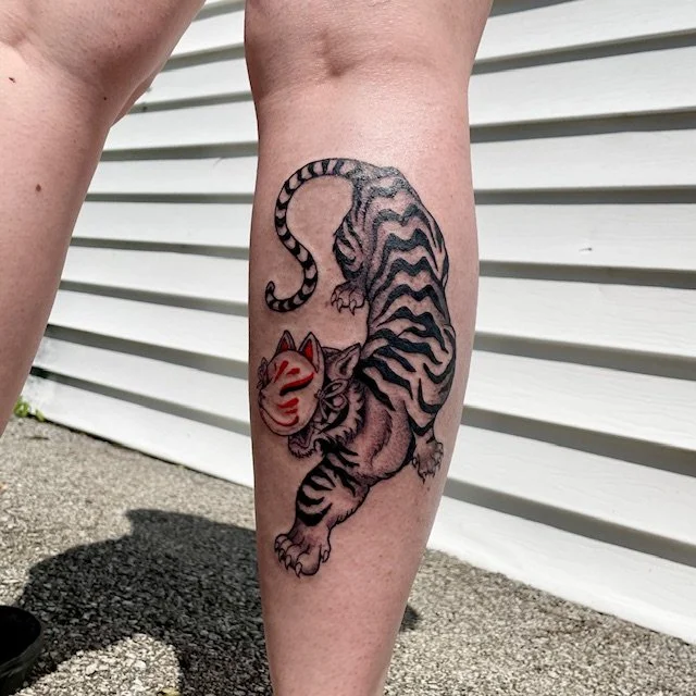 Tattoo of a tiger with a traditional Japanese mask on its face on a person’s calf outside next to a white siding wall.