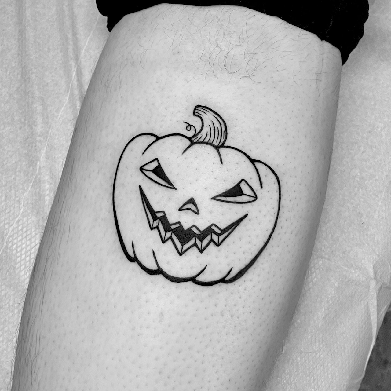 Black and white tattoo of a carved pumpkin with a mischievous face, jagged mouth, and narrowed eyes, with a small curly stem on top, on a person's upper arm.
