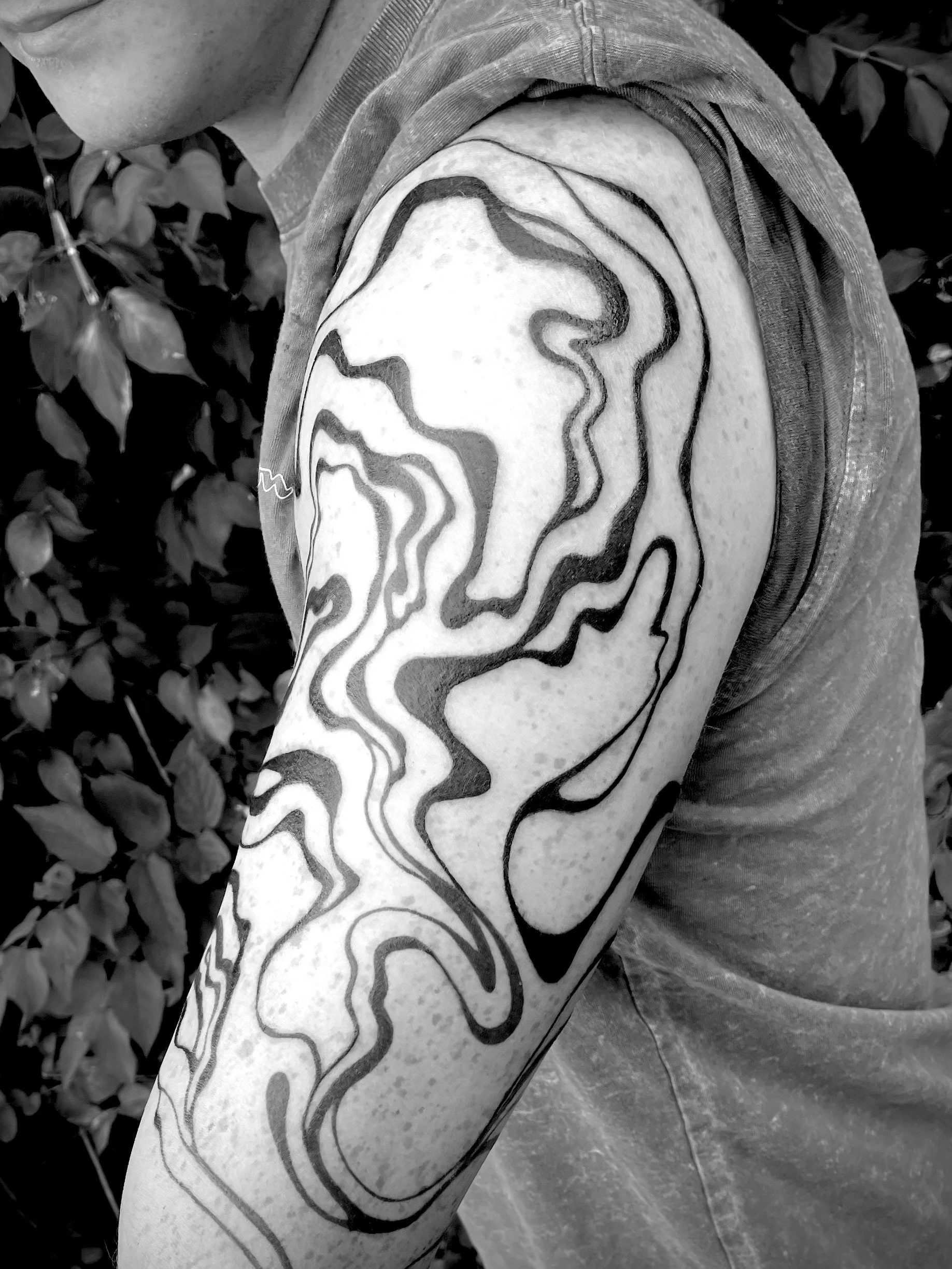 A person's upper arm features a tattoo with abstract, flowing black lines on the shoulder, with leafy plants in the background.